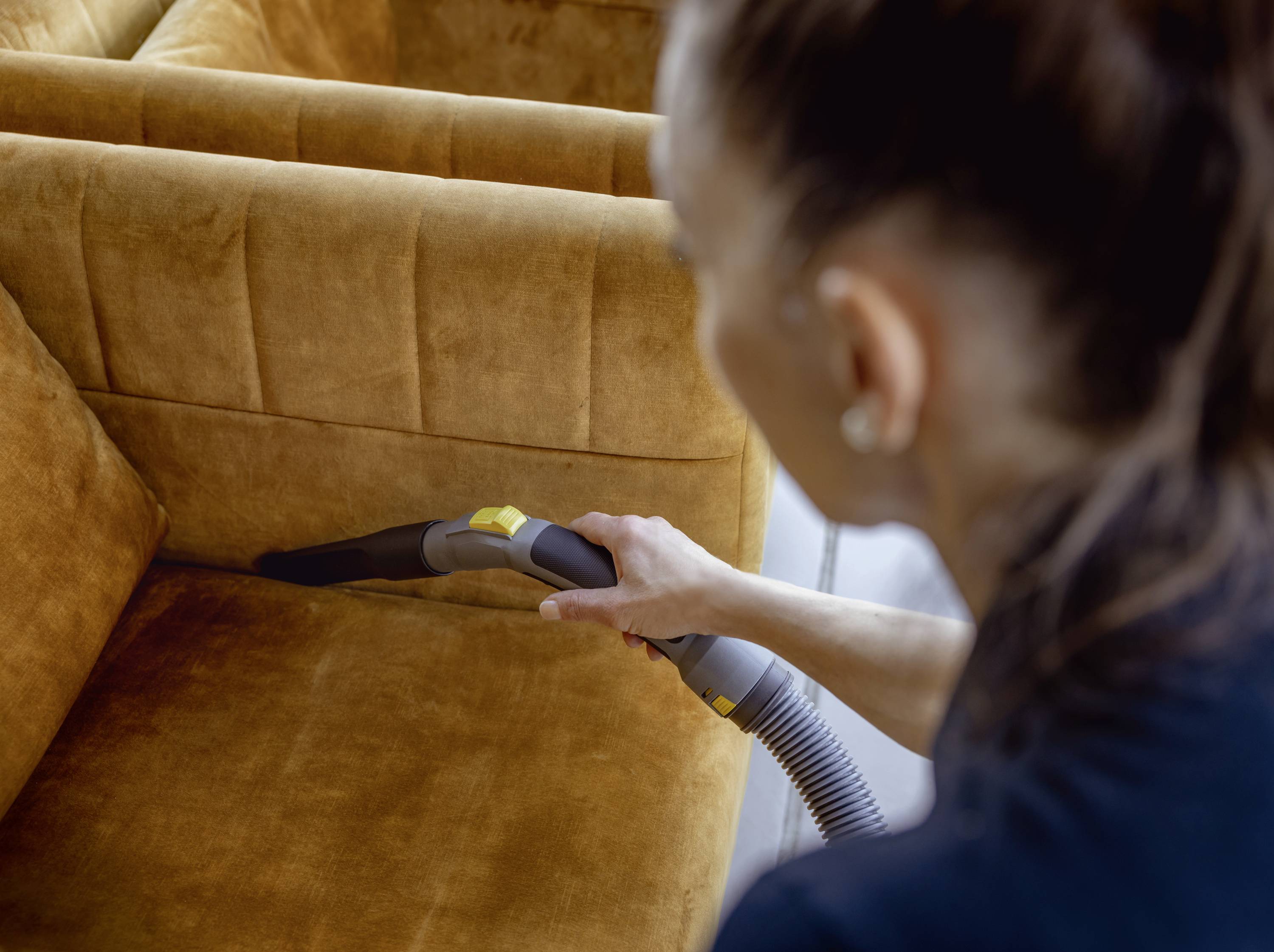 Een persoon zuigt de bank schoon met een stofzuiger. Focus op het reinigen van de naden in de mostersgele stoffen bank.
