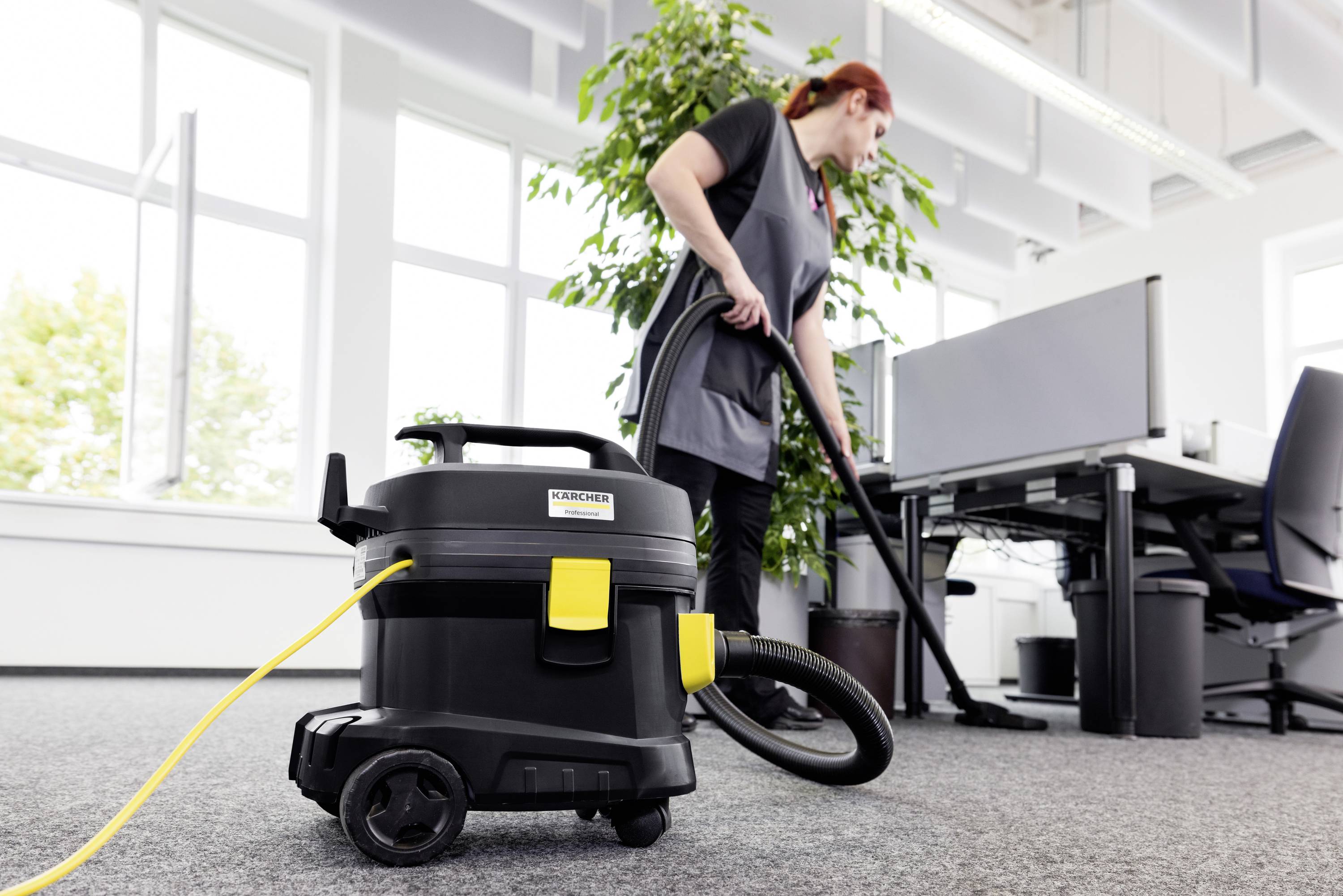 Een vrouw zuigt een kantoor met een industriële stofzuiger. Op de achtergrond staan bureaus en een grote groene plantenpot.