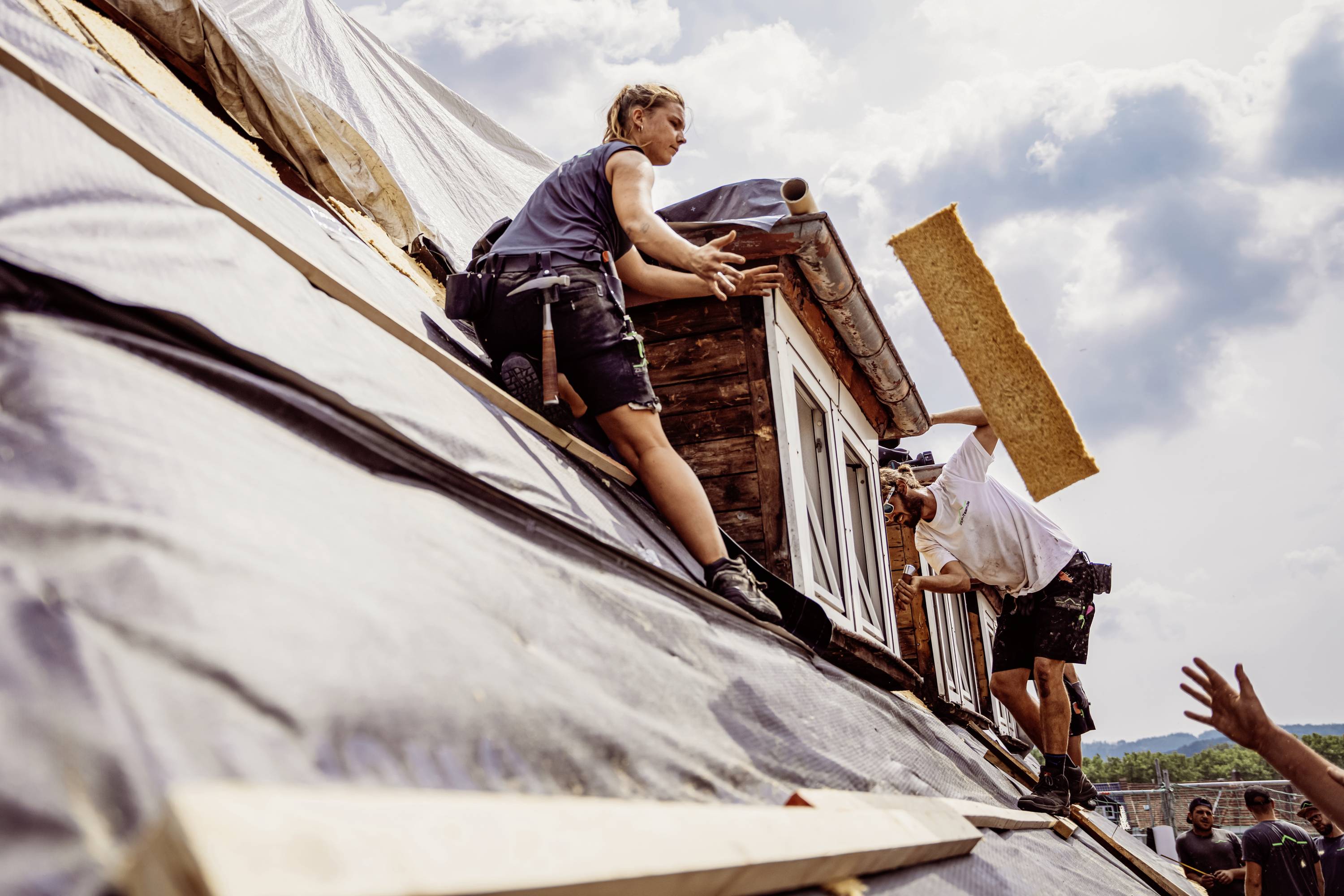 Twee personen werken op het schuine dak van een huis, een van hen houdt een houten plank vast, de ander een zaag, bij zonnig weer.
