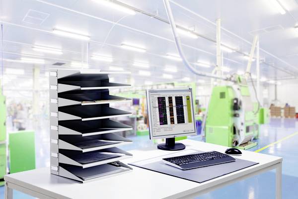 In een helder fabrieksruimte staat een bureau met computer, toetsenbord en documentenhouder; op de achtergrond zijn machines zichtbaar.