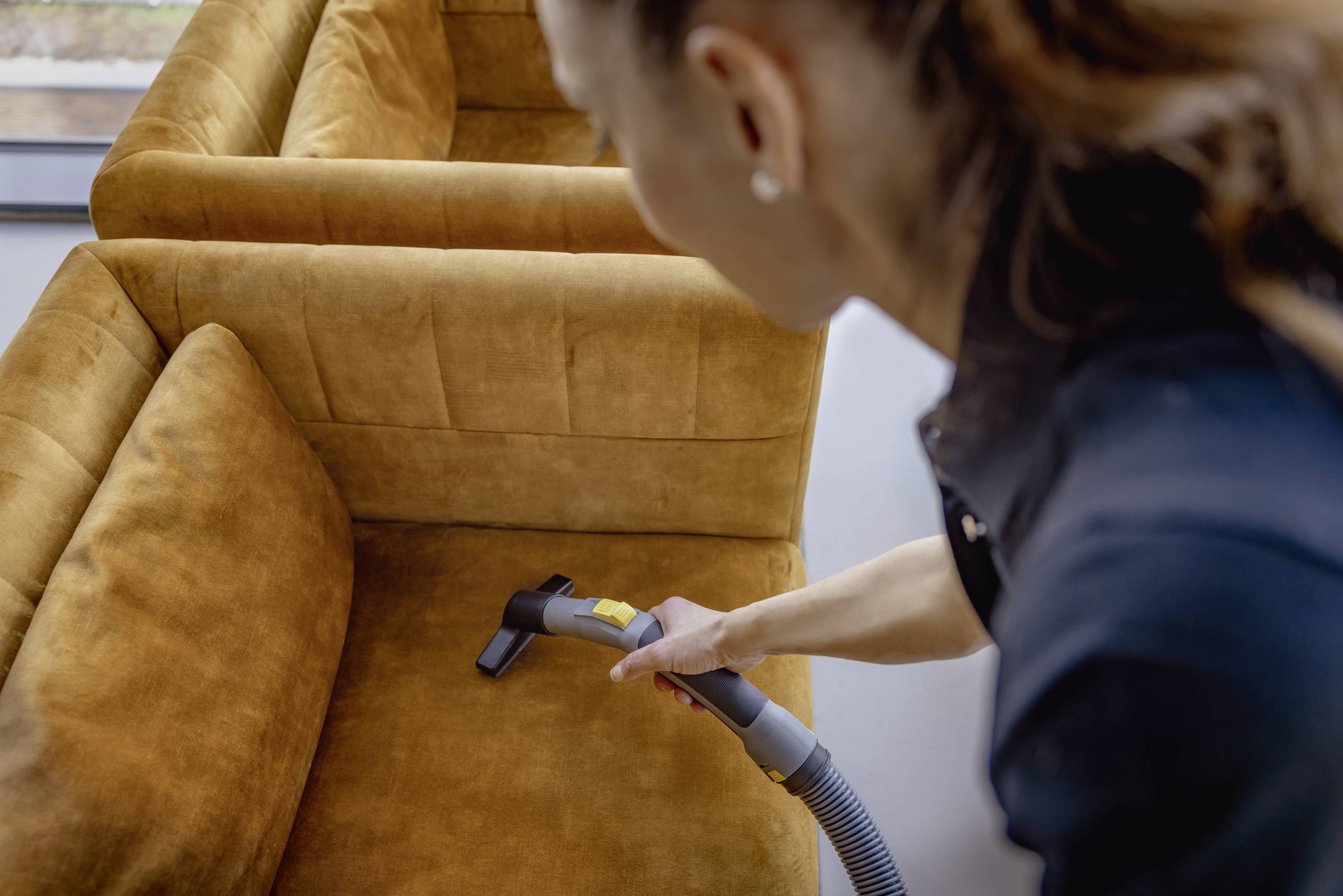Een persoon zuigt een bruine bank schoon met een meubelzuigmond. De focus ligt op het reinigen van het zitmeubel.