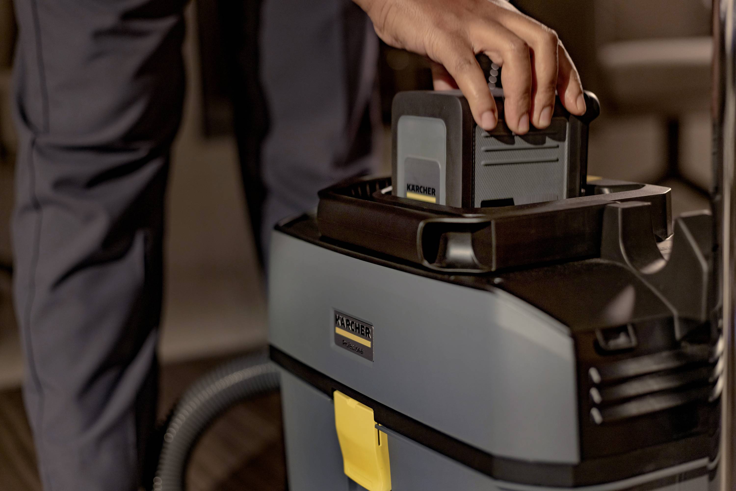 Een persoon plaatst een batterij in een industriële stofzuiger. De stofzuiger is grijs met een geel element en heeft een robuuste constructie.