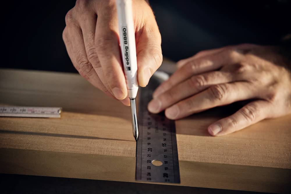 Een persoon tekent met een pen op een houten bord, ondersteund door een metalen liniaal.