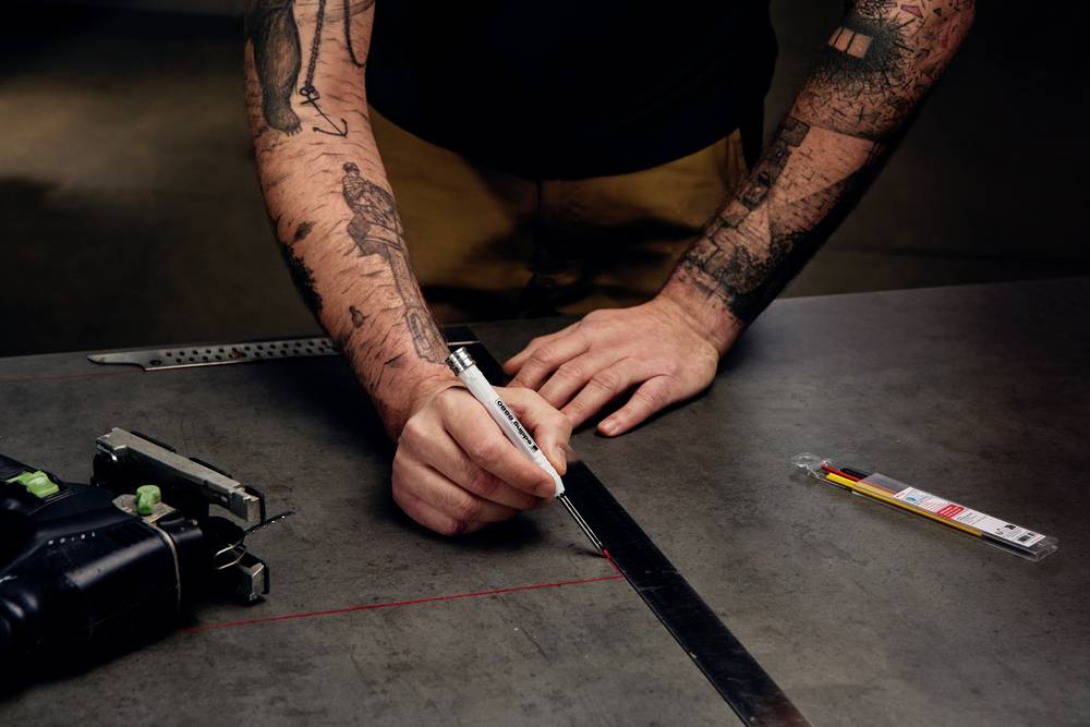 Een getatoeëerde persoon meet en tekent een lijn op een metalen oppervlak met een liniaal en een pen. Gereedschap links in de afbeelding.