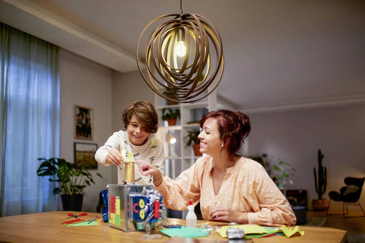 Een kind en een vrouw knutselen samen aan een tafel, omringd door papier en knutselspullen. Beiden zien er blij en geconcentreerd uit.