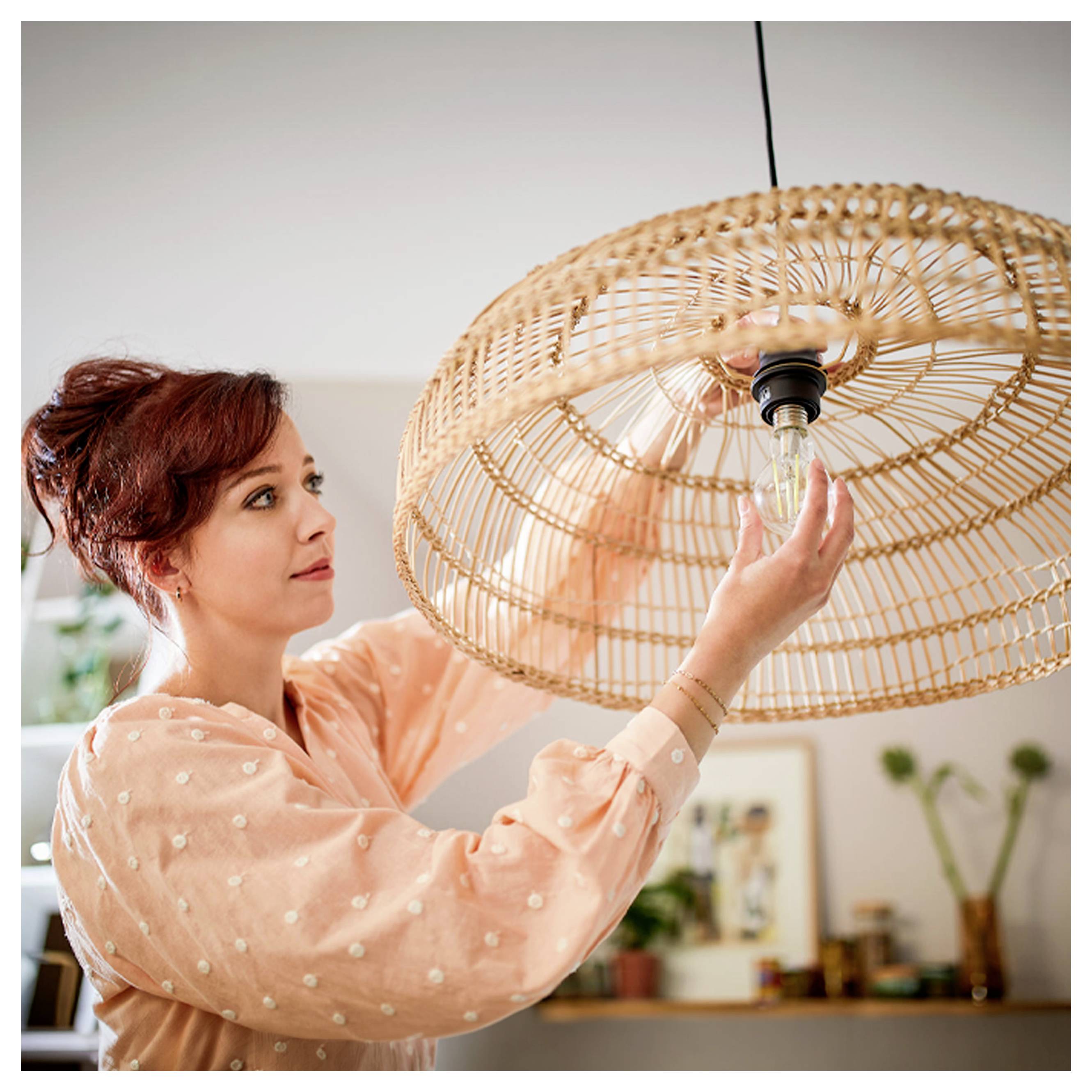 Vrouw in een perzikkleurige blouse installeert een gloeilamp in een gevlochten hanglamp binnenshuis.