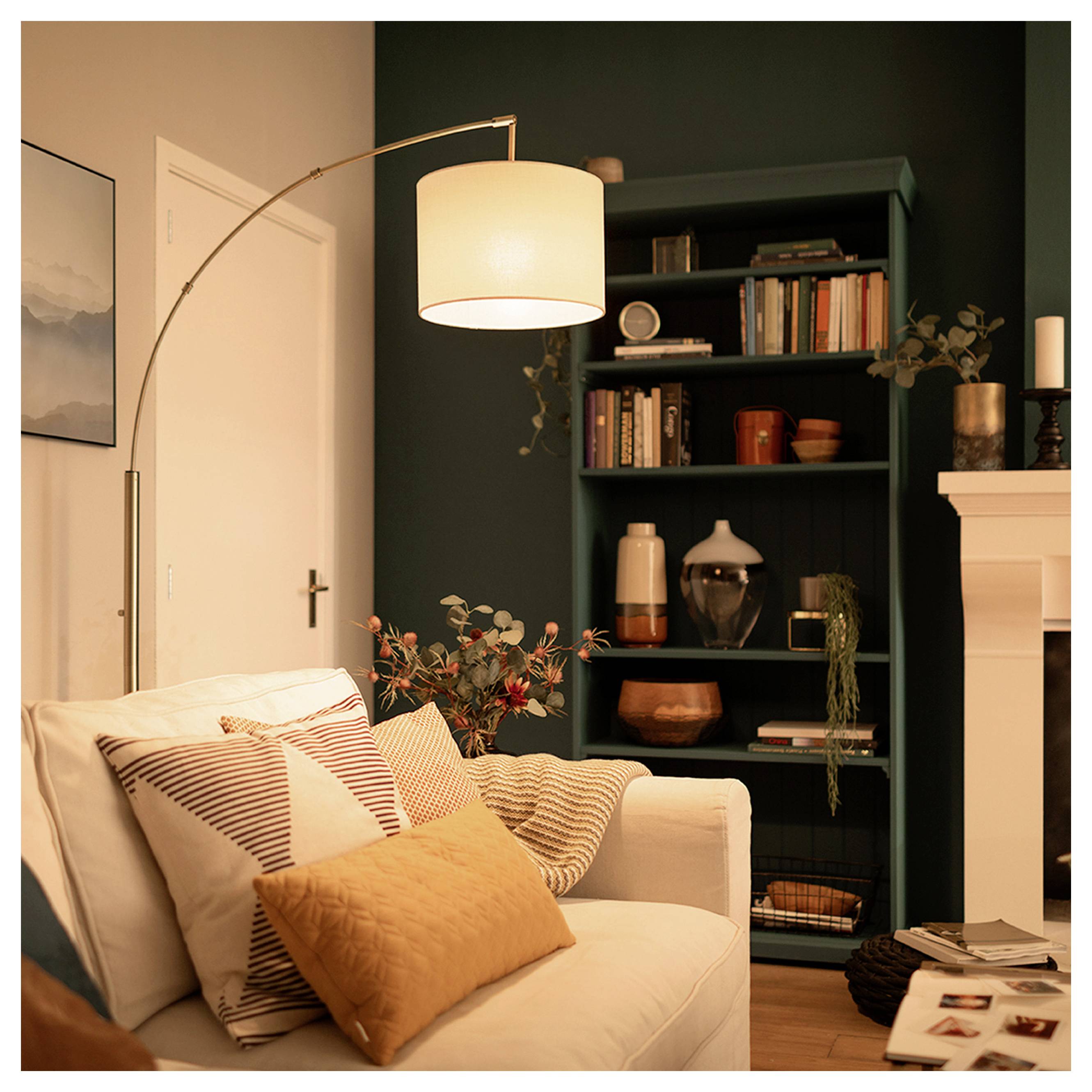 Gezellige woonkamer met een witte bank, gemusterde kussens, staande lamp, donkere boekenkast en schoorsteenmantel.