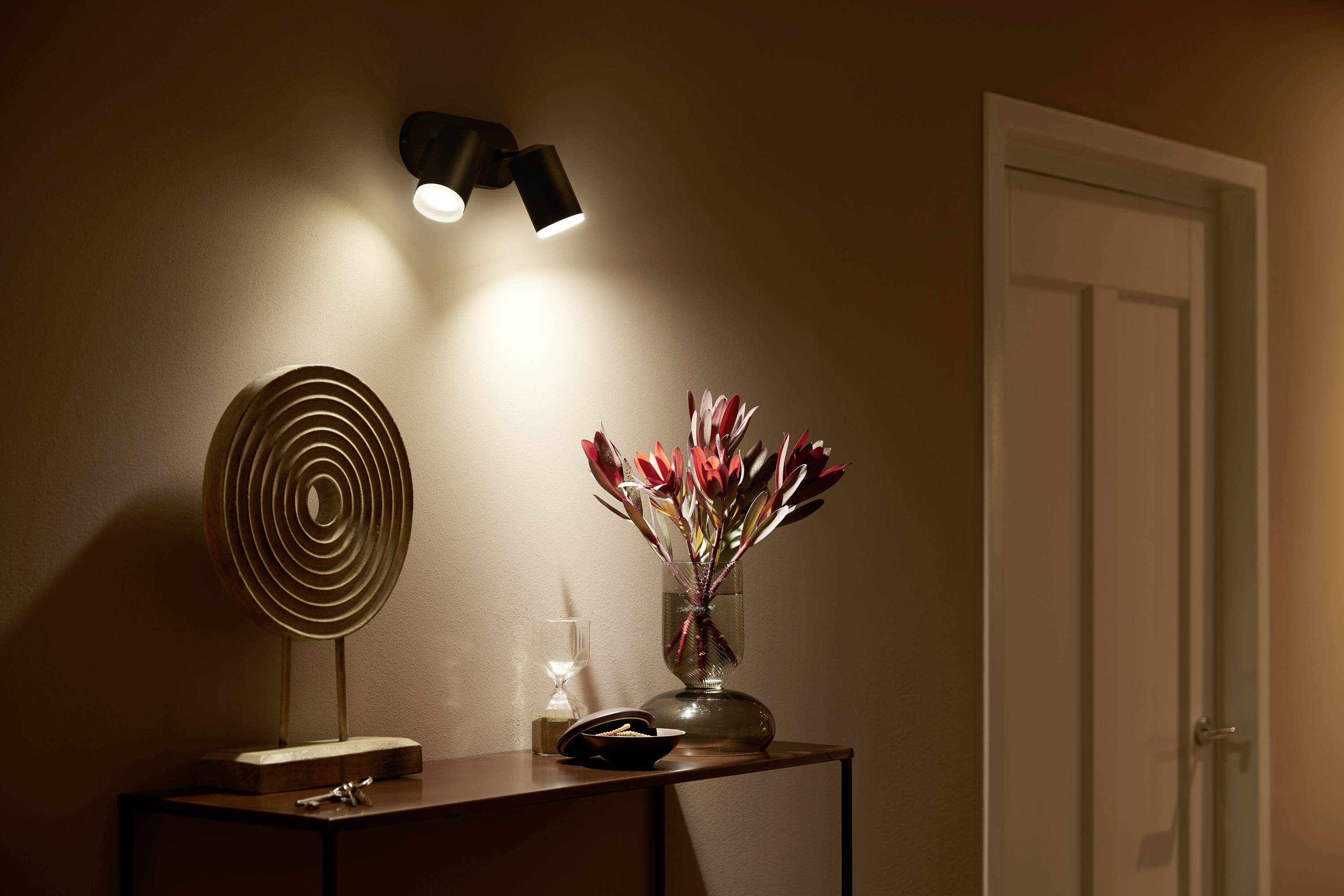 Een sfeervol verlichte gang met een moderne sculptuur en een vaas met rode bloemen op een dressoir.