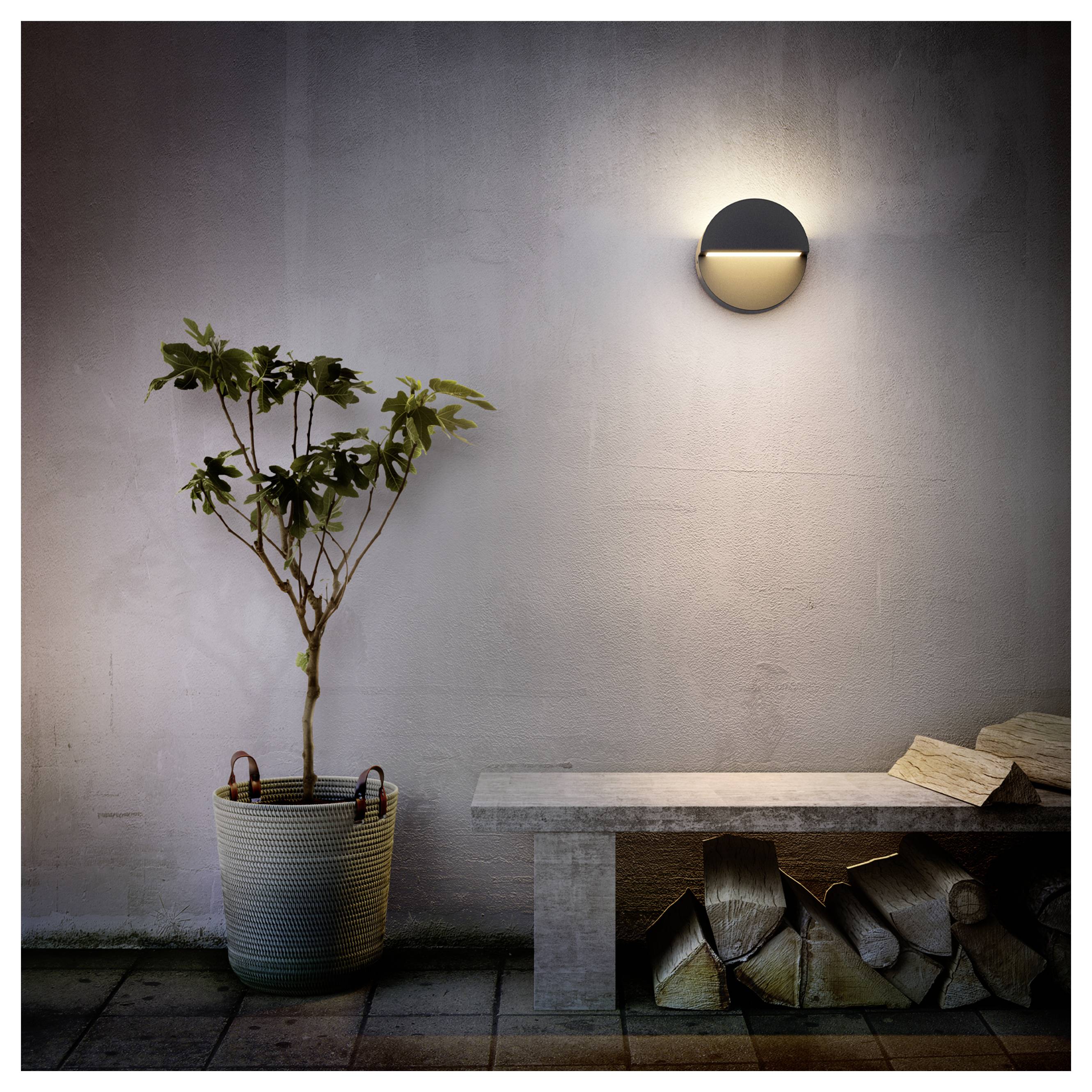 Minimalistische patio met een potplant boom, stenen bank, gestapeld brandhout en een verlichte wandlamp tegen een eenvoudige muur.