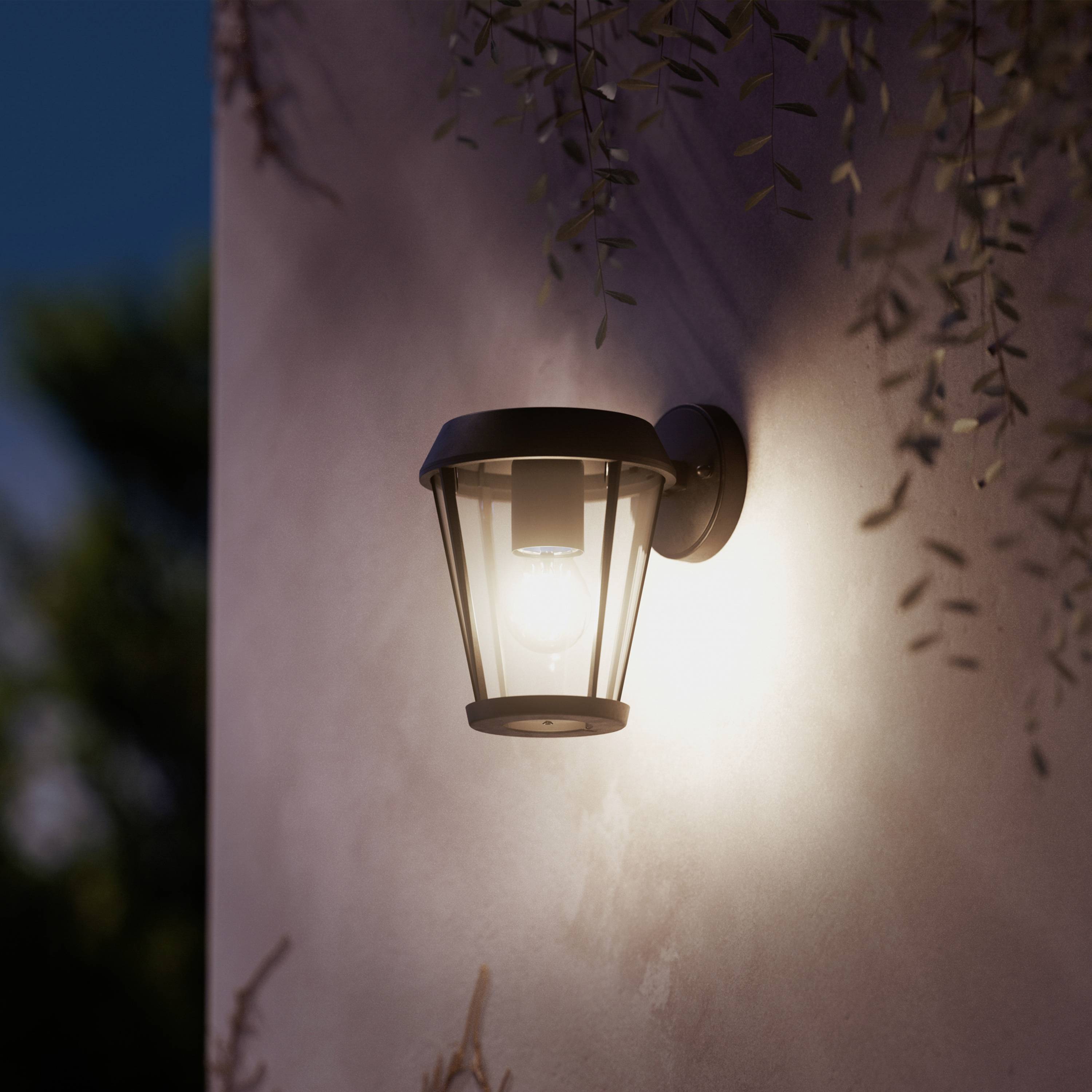 Buitenlamp aan een muur verlicht zacht de omgeving in de schemering, omgeven door planten.