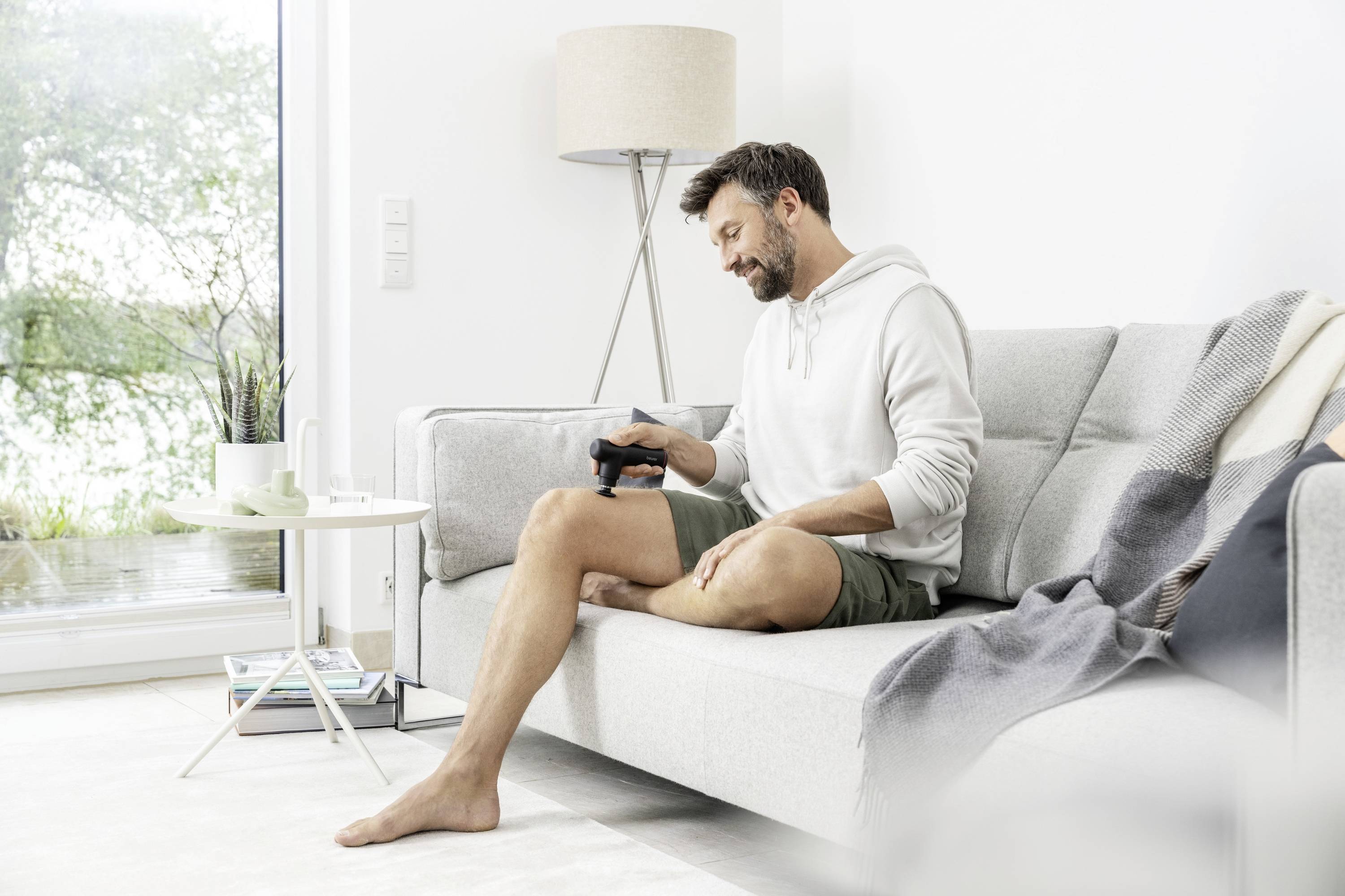 Een man zit op een bank in een lichte woonkamer en gebruikt een massageapparaat op zijn been. Naast hem staat een tafel met kopjes.