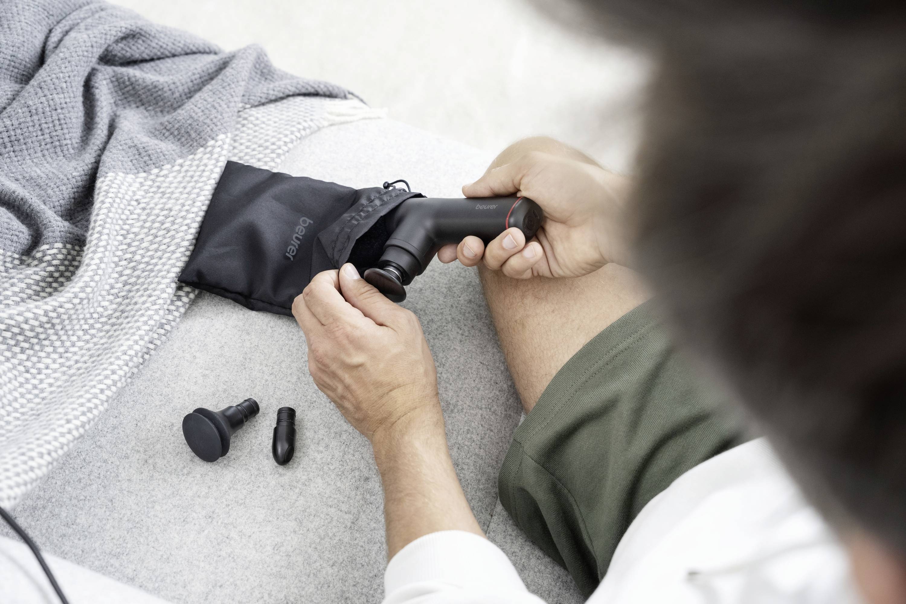 Een persoon haalt een massagepistool uit een zwart etui. Drie verschillende opzetstukken liggen ernaast.