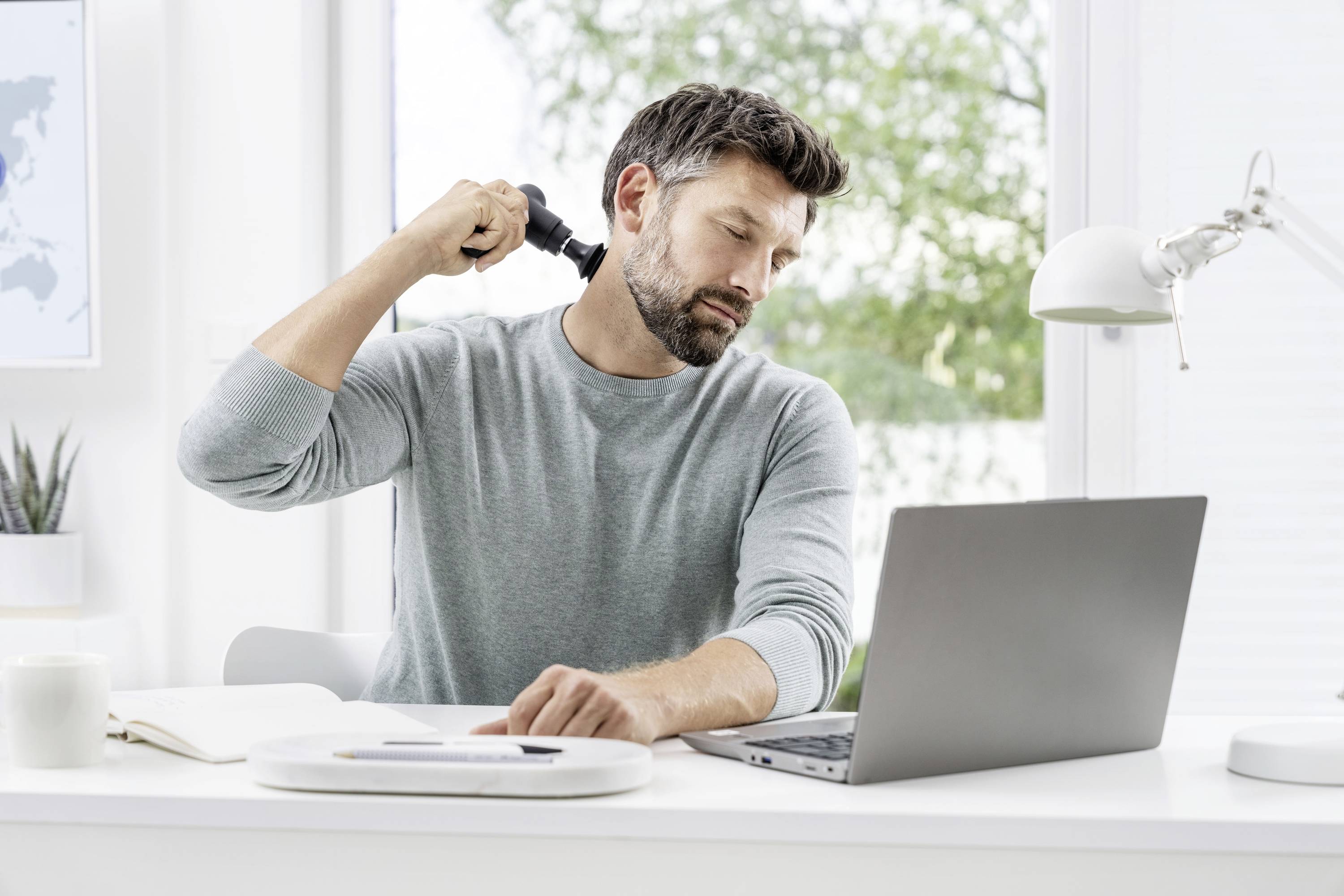 Een man in een lichtgrijze trui zit aan een bureau met een laptop en gebruikt een handmassageapparaat op zijn nek.