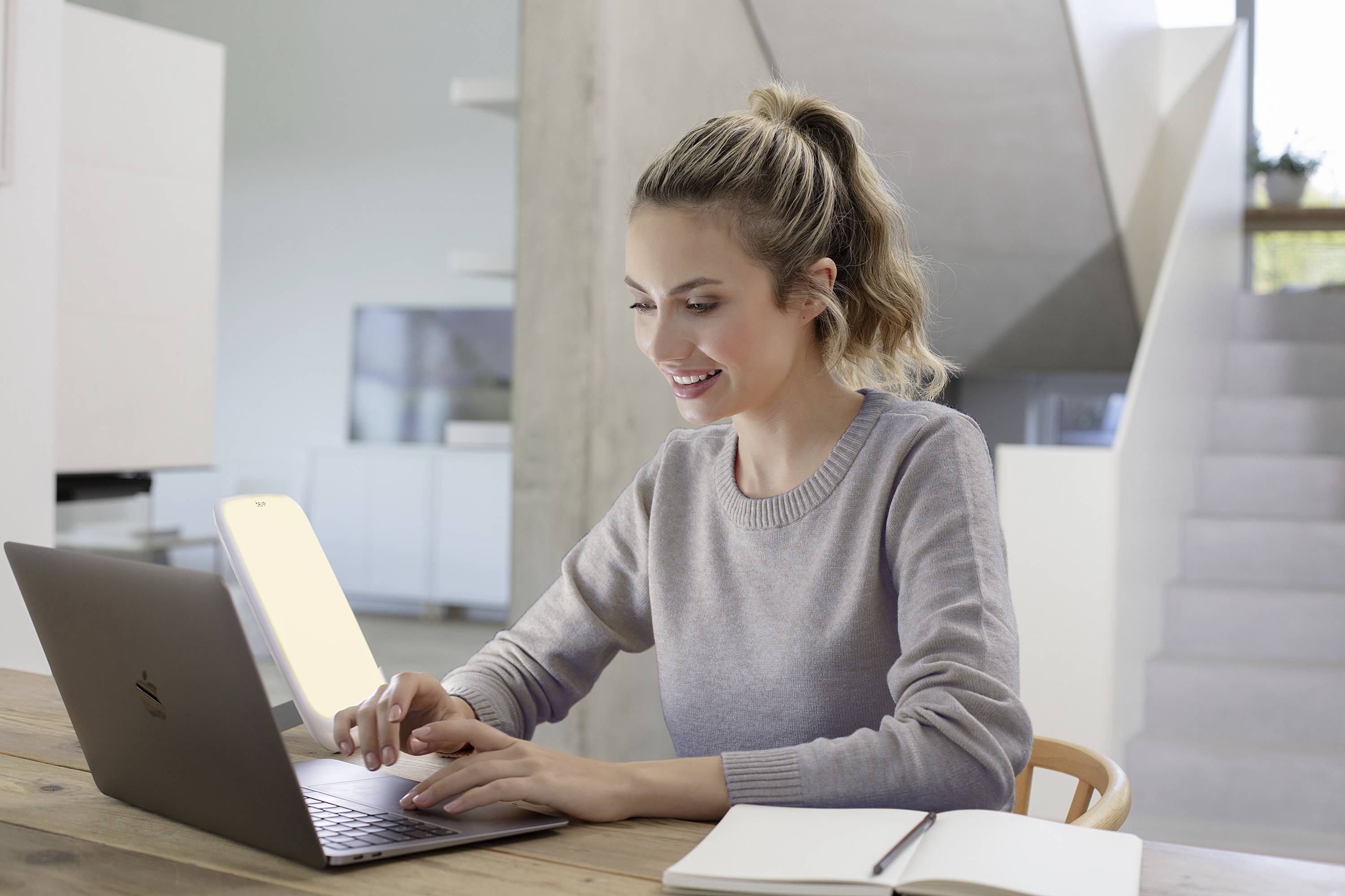 Een lachende vrouw zit aan een tafel en typt op een laptop. Naast haar ligt een notitieboek. Op de achtergrond zijn moderne meubels zichtbaar.