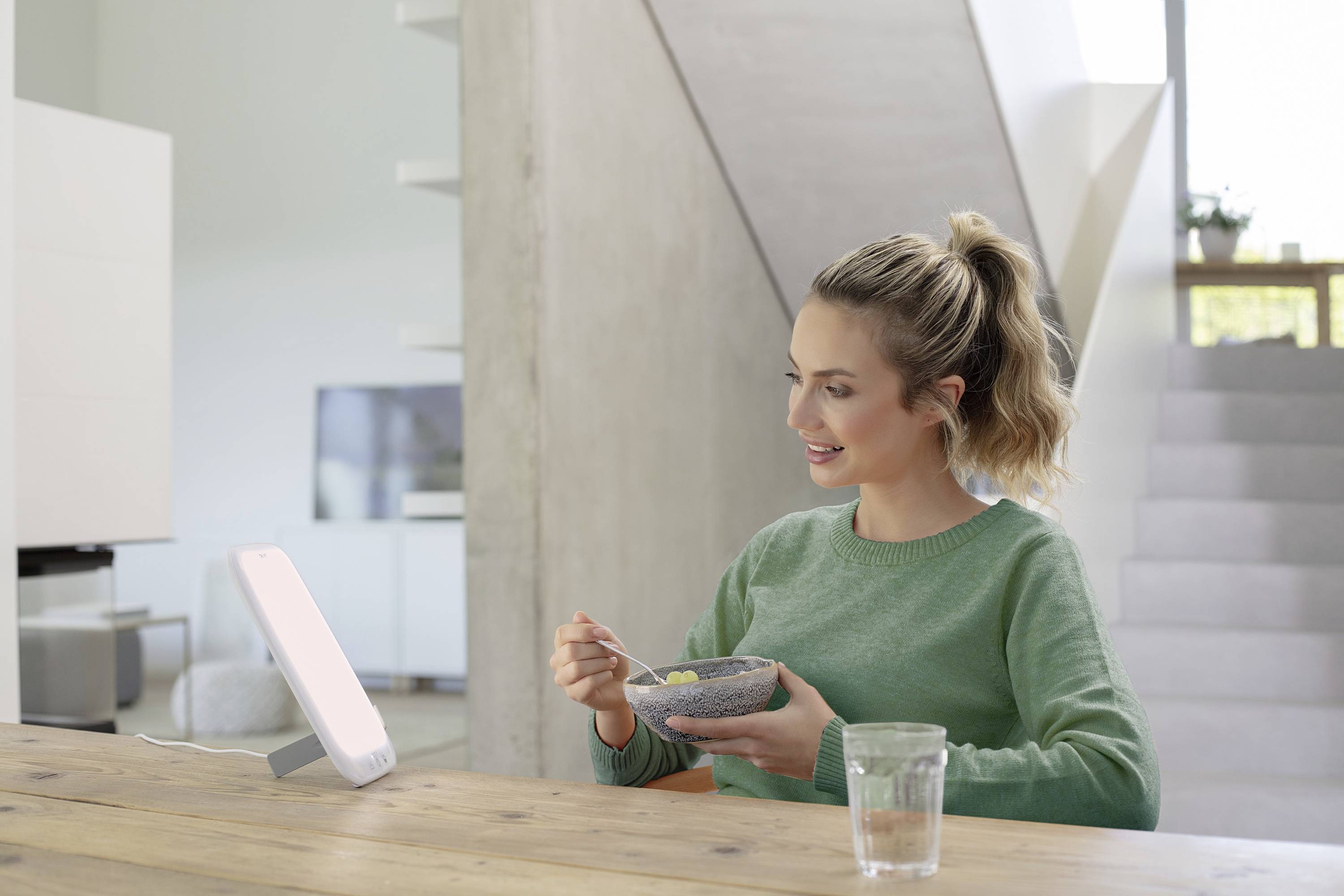 Een vrouw zit aan tafel, eet uit een kom en kijkt naar een heldere daglichtlamp. Op de achtergrond is een modern woonvertrek.