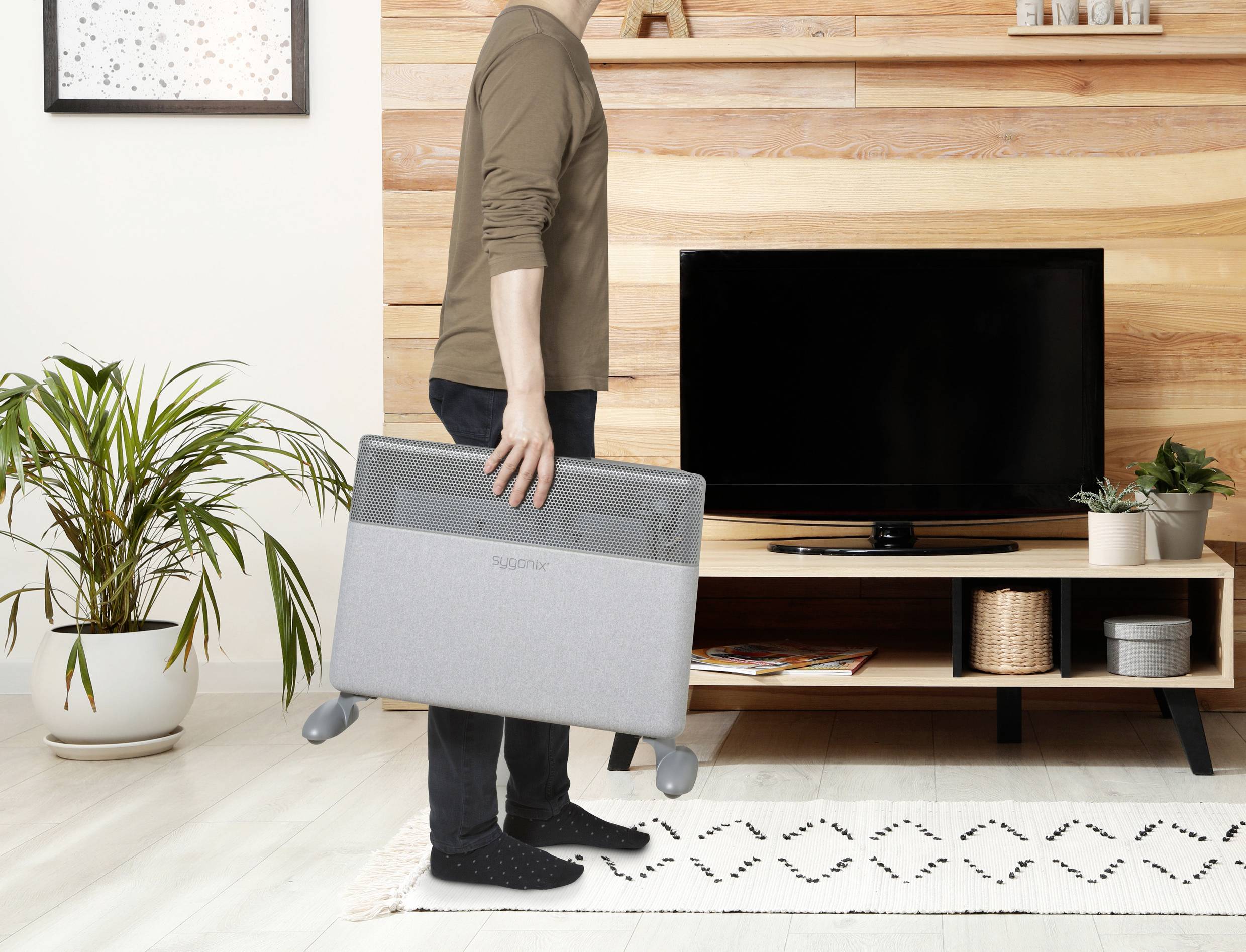 Een persoon draagt een grijze elektrische radiator in een woonkamer met houten wanden, een zwarte televisie en planten.