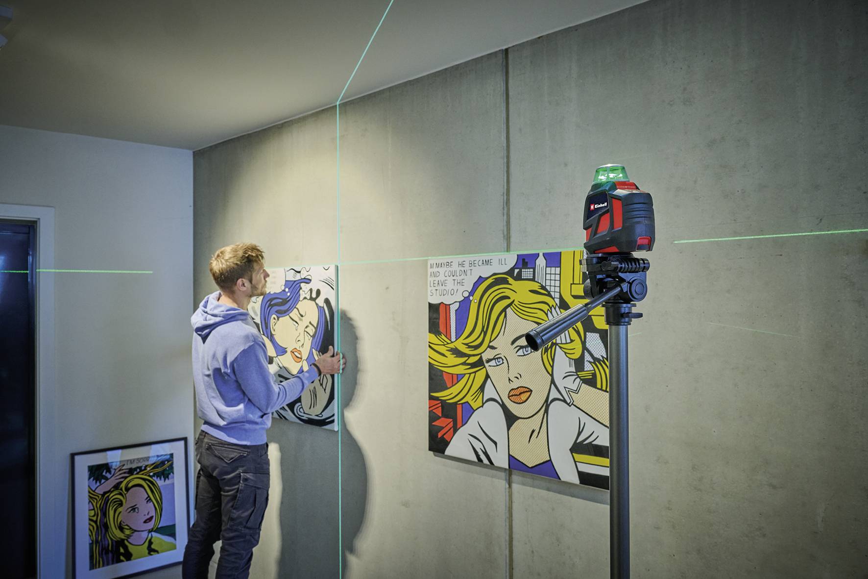 Een man hangt een schilderij aan een muur op, ondersteund door een groen laserapparaat voor uitlijning. Op de achtergrond zijn nog meer schilderijen zichtbaar.