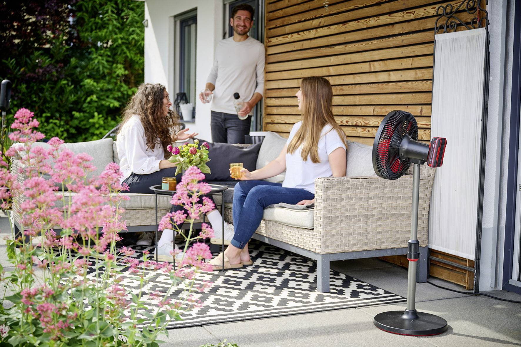 Een groep van drie personen zit en staat op een terras. Twee vrouwen zitten op een bank, terwijl een man staat. Er zijn planten op de voorgrond.