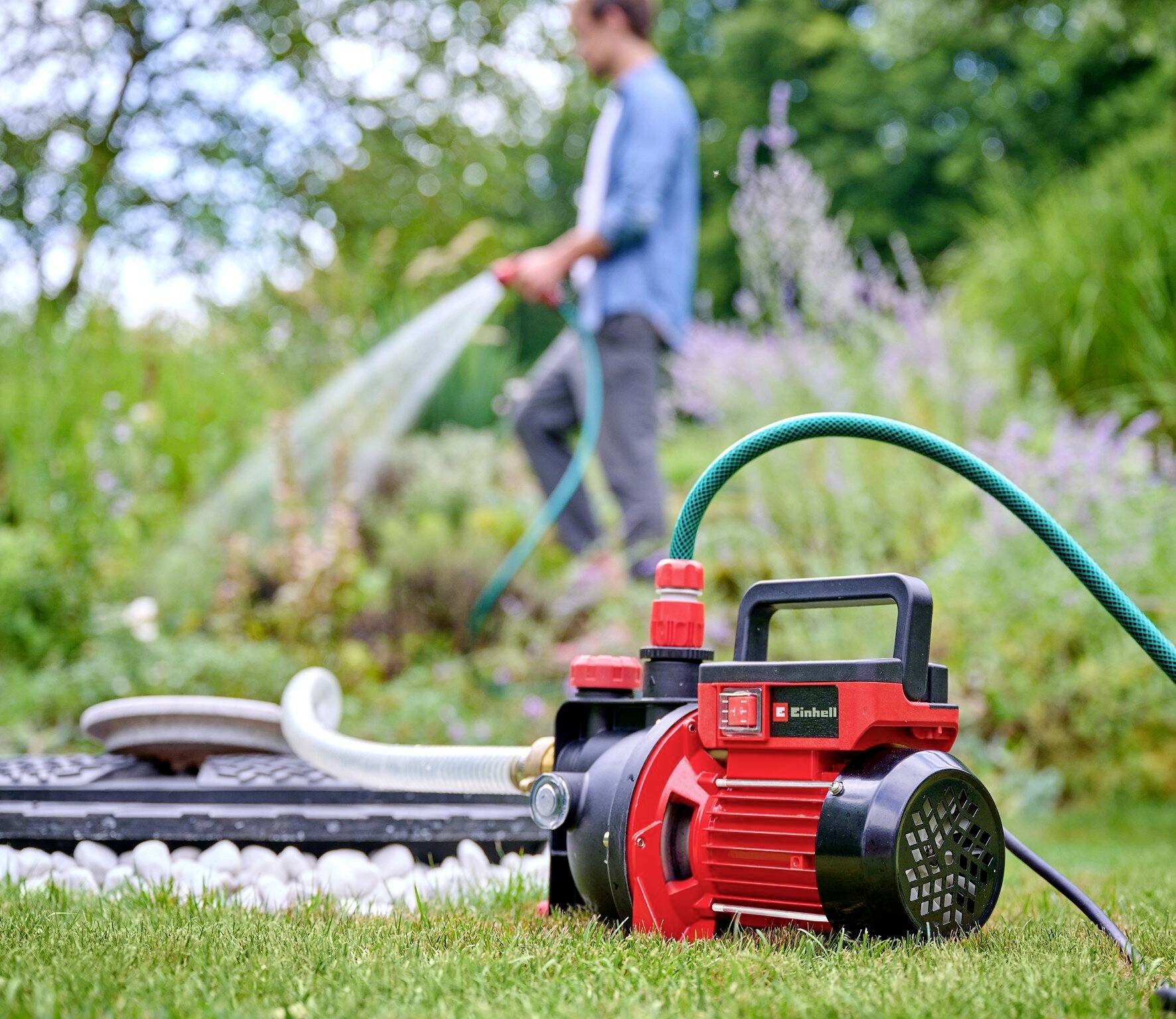 Een persoon besproeit planten op de achtergrond met een tuinslang. Op de voorgrond staat een rode elektrische tuinpomp op het gazon.
