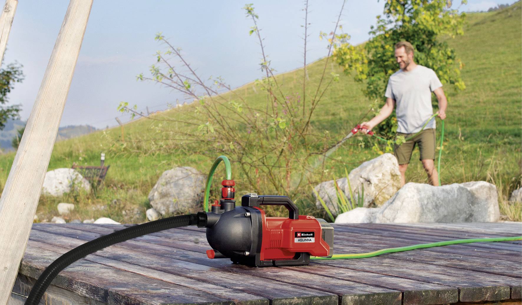 Een persoon besproeit planten in een tuin met een tuinslang. Een waterpomp is prominent in de voorgrond zichtbaar.