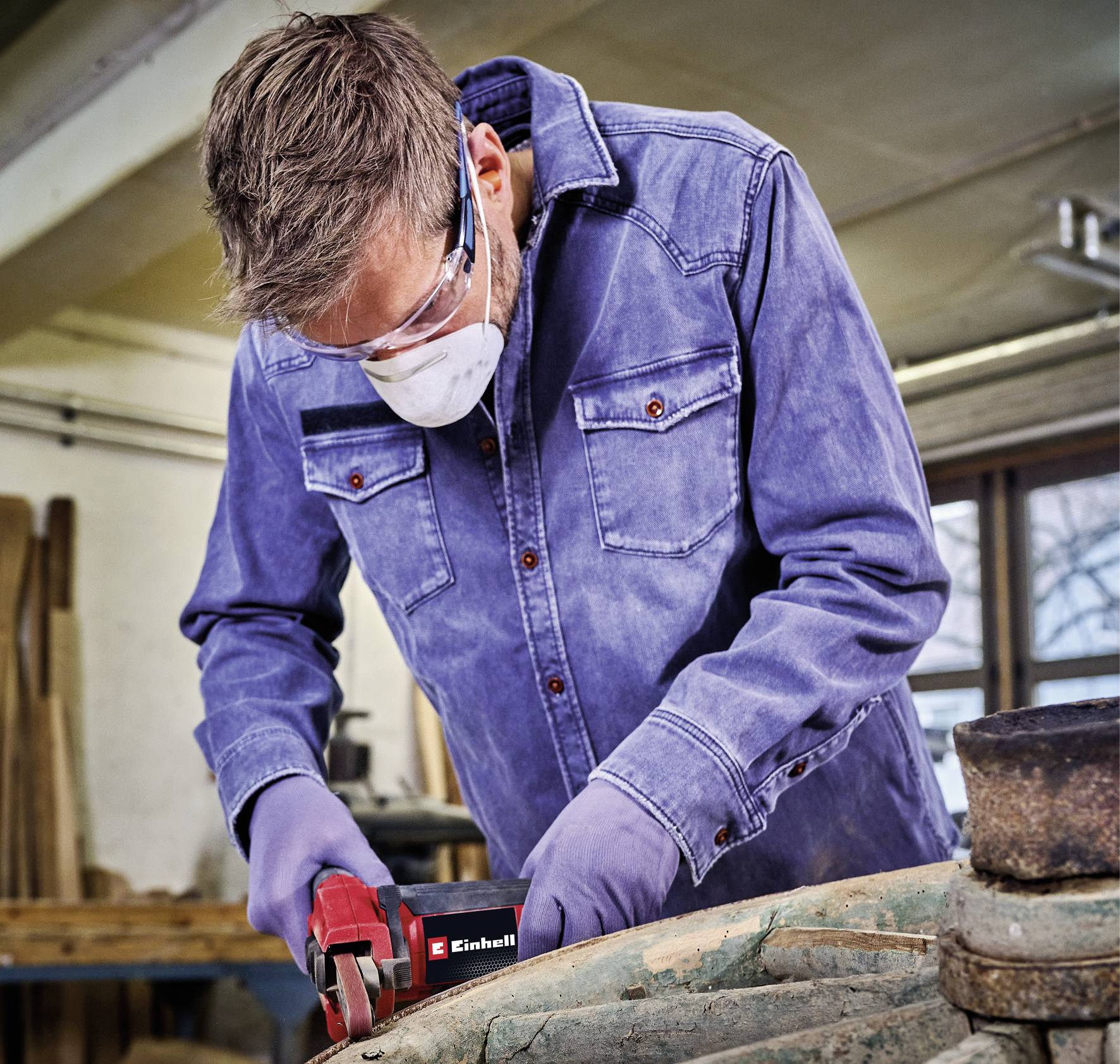 Een man met veiligheidsbril en masker werkt in een werkplaats met een elektrisch gereedschap aan een stuk hout.