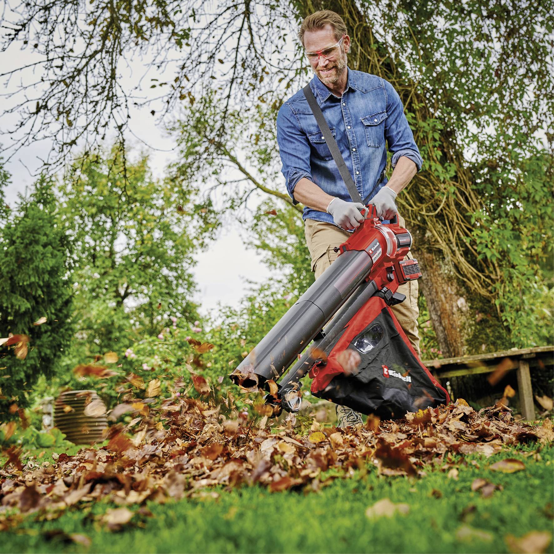 Man in spijkerhemd gebruikt bladblazer in tuin, blaast gekleurde herfstbladeren van gazon; bomen en struiken op de achtergrond.