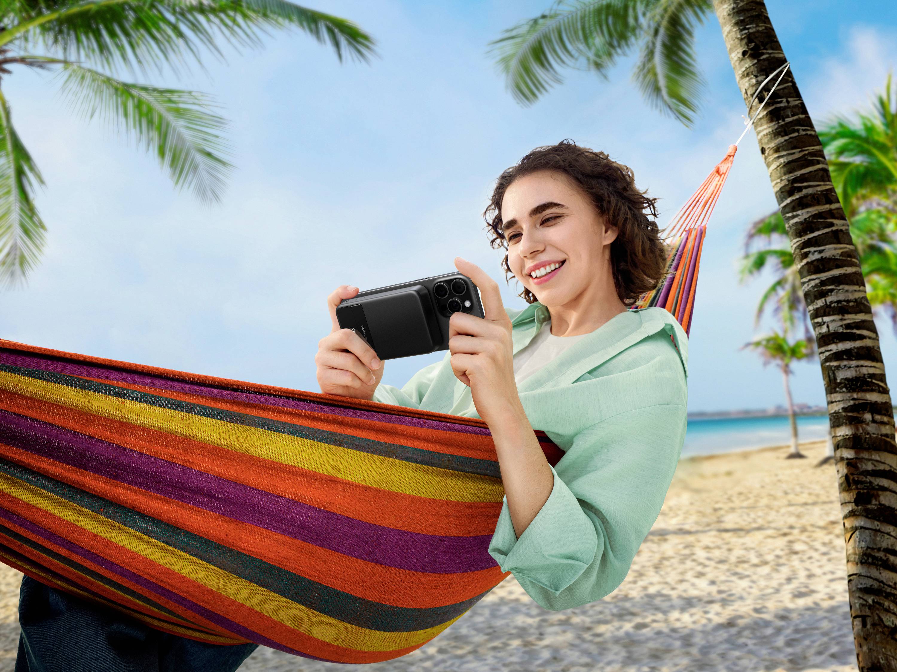 Een vrouw ligt in een kleurrijke hangmat op het strand en glimlacht terwijl ze naar haar smartphone kijkt; palmbomen op de achtergrond.