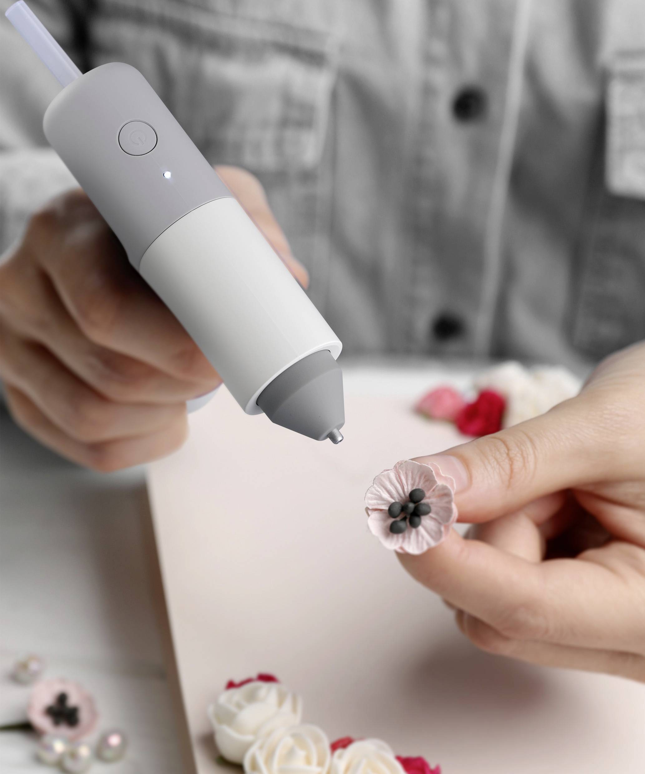 Een persoon houdt een lijmpistool vast en plakt een kleine, roze kunstbloem op een vel papier. Nog enkele bloemen liggen ernaast.