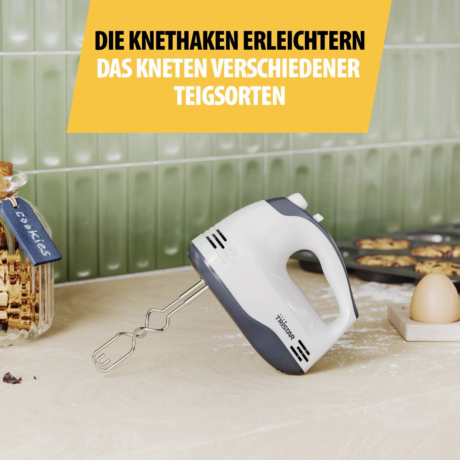 Een handmixer met deeghaken staat op een aanrecht. Op de achtergrond zijn koekjes en een eierhouder te zien.