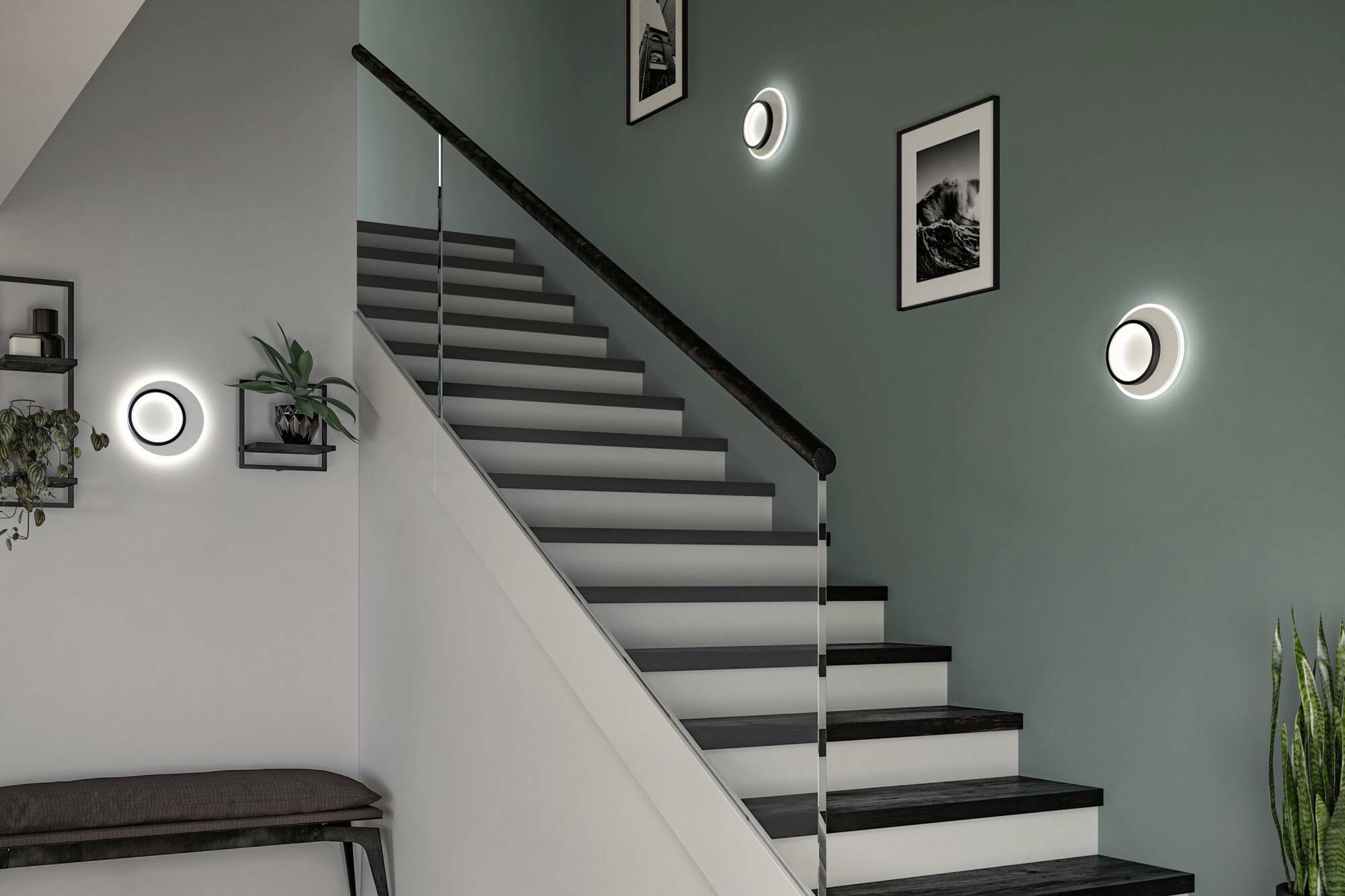 Een trap met zwarte leuningen leidt naar boven. Aan de muur hangen foto's en ronde, verlichte lampen. Planten staan naast de trap.