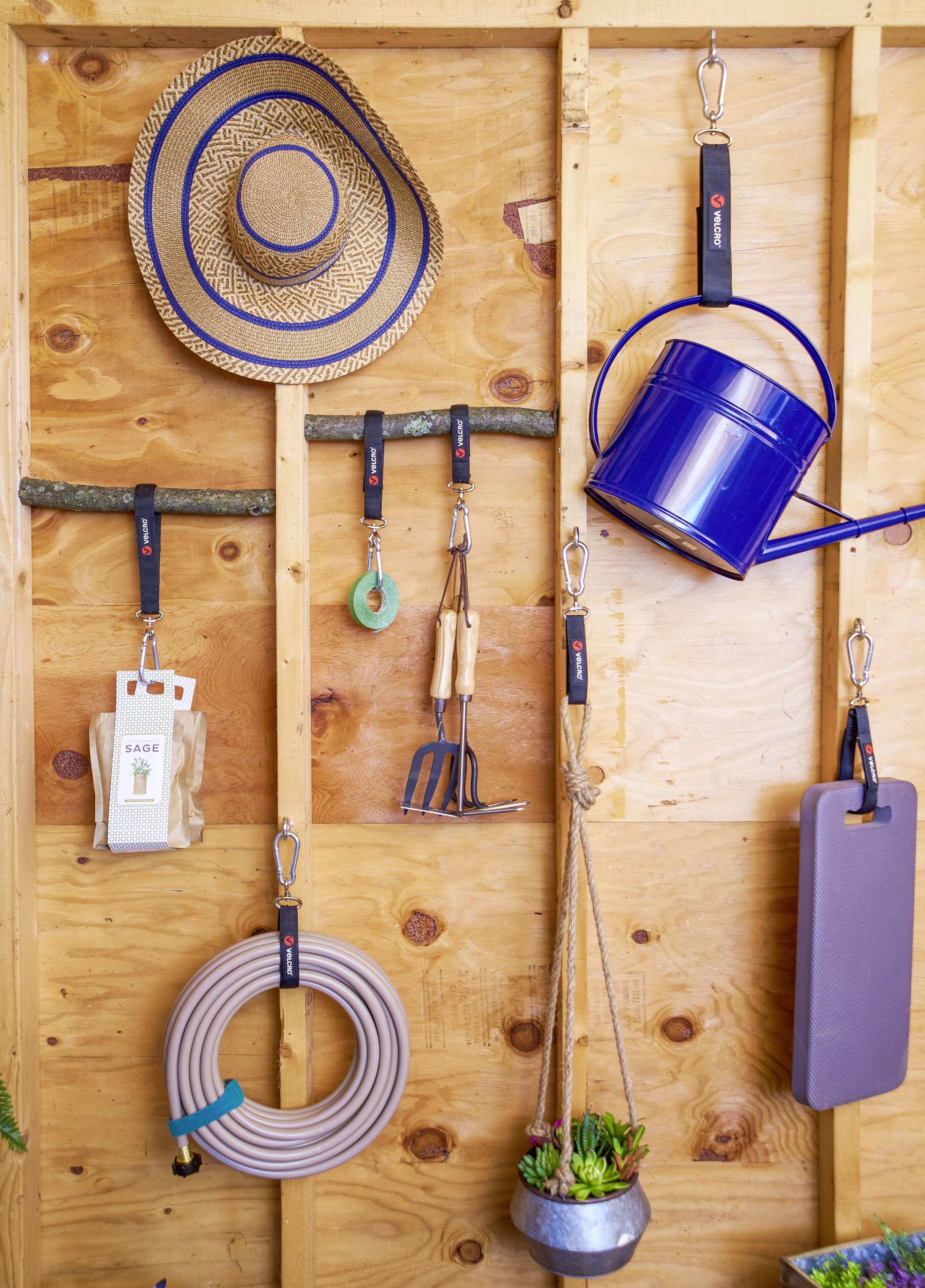 Tuingereedschappen en toebehoren hangen netjes aan een houten wand, daaronder een hoed, een gieter, tuingereedschappen en een tuinslang.