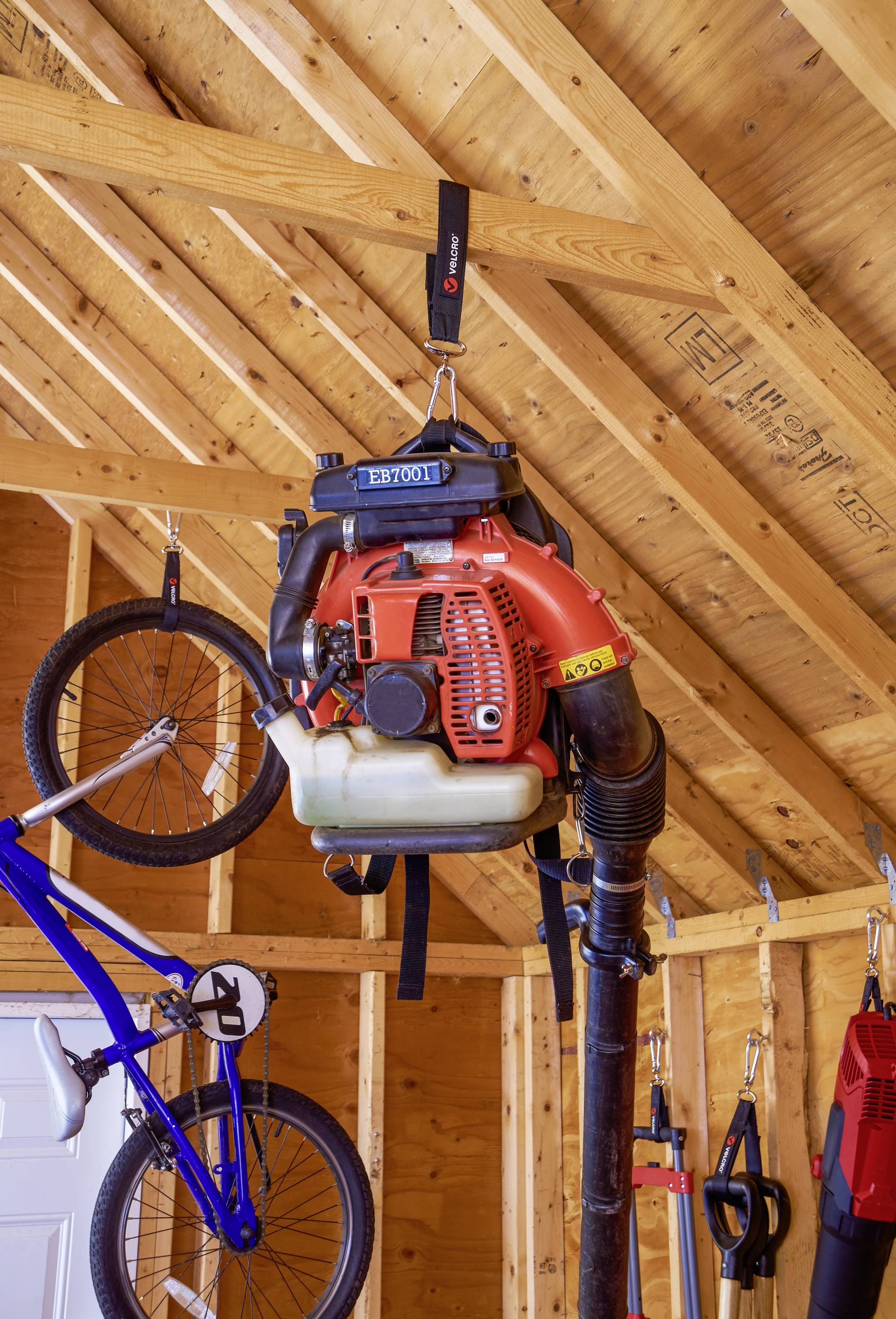 Een rode bladblazer hangt aan een houten frame in een garage. Eronder staat een blauwe fiets en tuingereedschap.