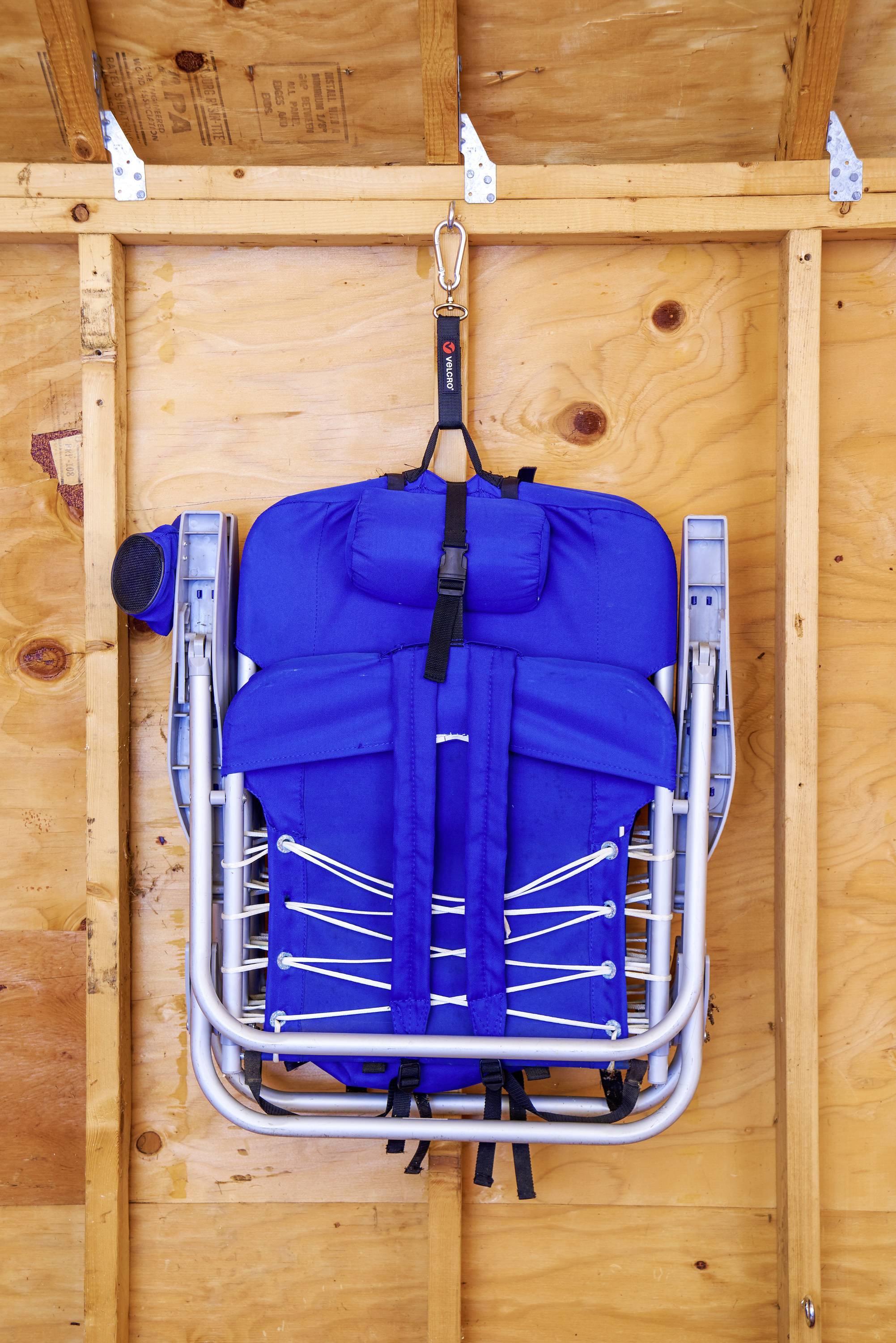 Blauwe opvouwbare stoel hangt aan een houten wand in een schuur. De stoel is compact ingeklapt en met een riem vastgemaakt.