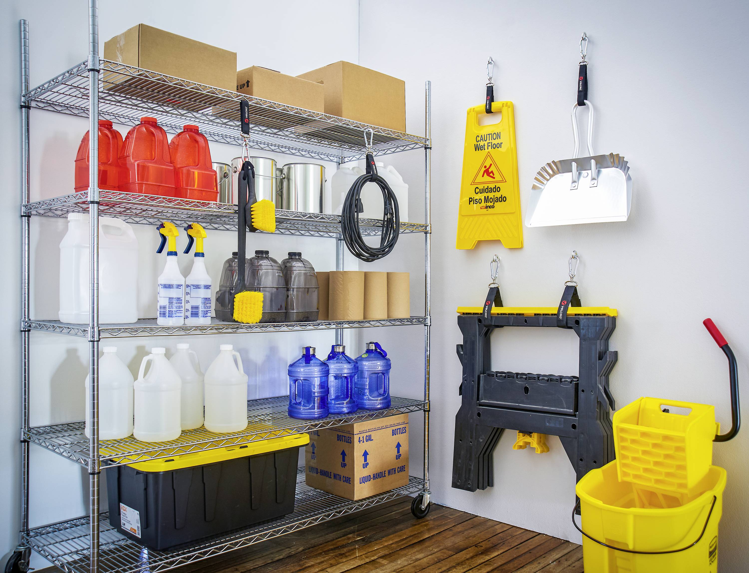 Een opslagruimte met rekken vol schoonmaakmiddelen, zoals jerrycans, spuitflessen en emmers. Dweil en waarschuwingsbord aan de muur.