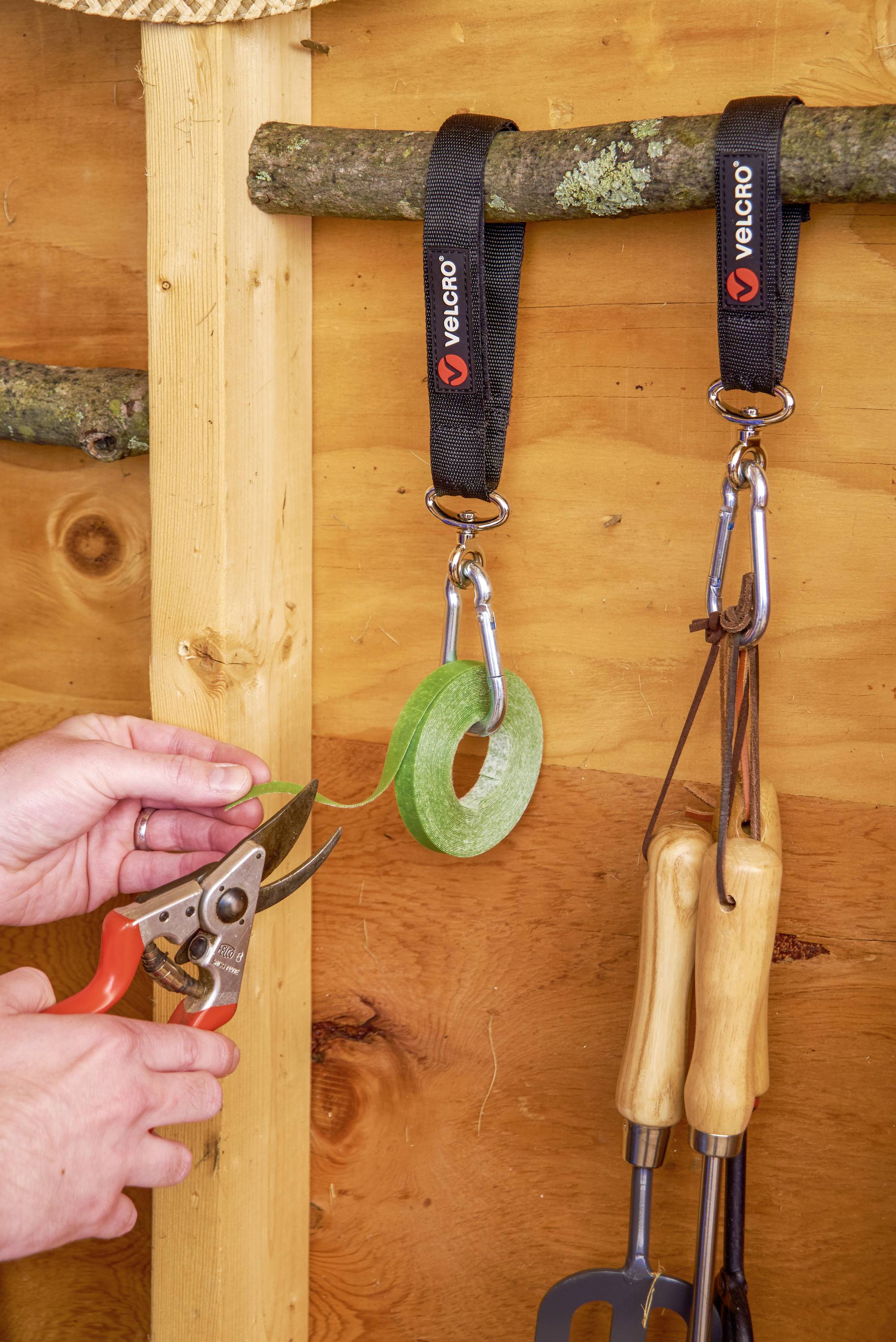 Een persoon knipt een plakband door met een schaar in een houten hut. Op de achtergrond zijn haken aan de muur bevestigd met tuingereedschappen.