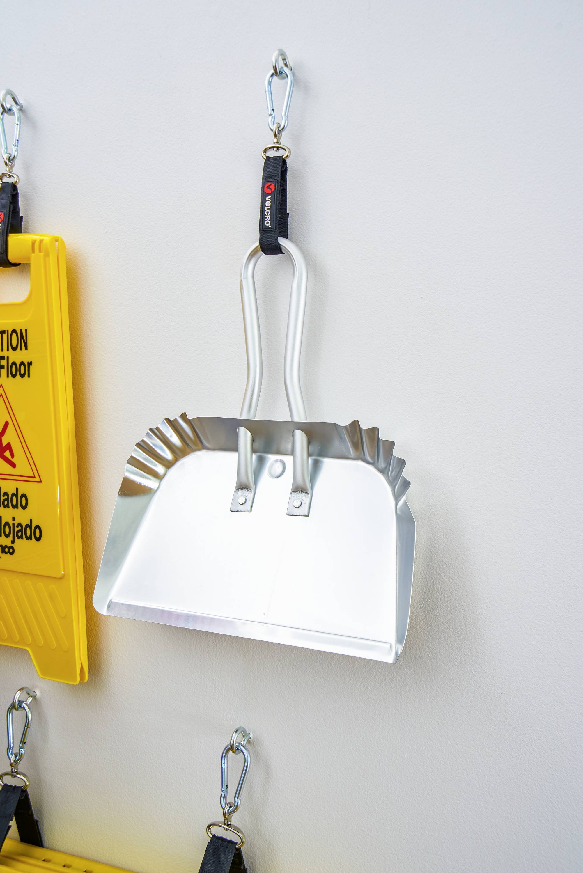 Zilveren metalen schep hangt aan de muur, ernaast een geel waarschuwingsbord 'Let op: natte vloer'.