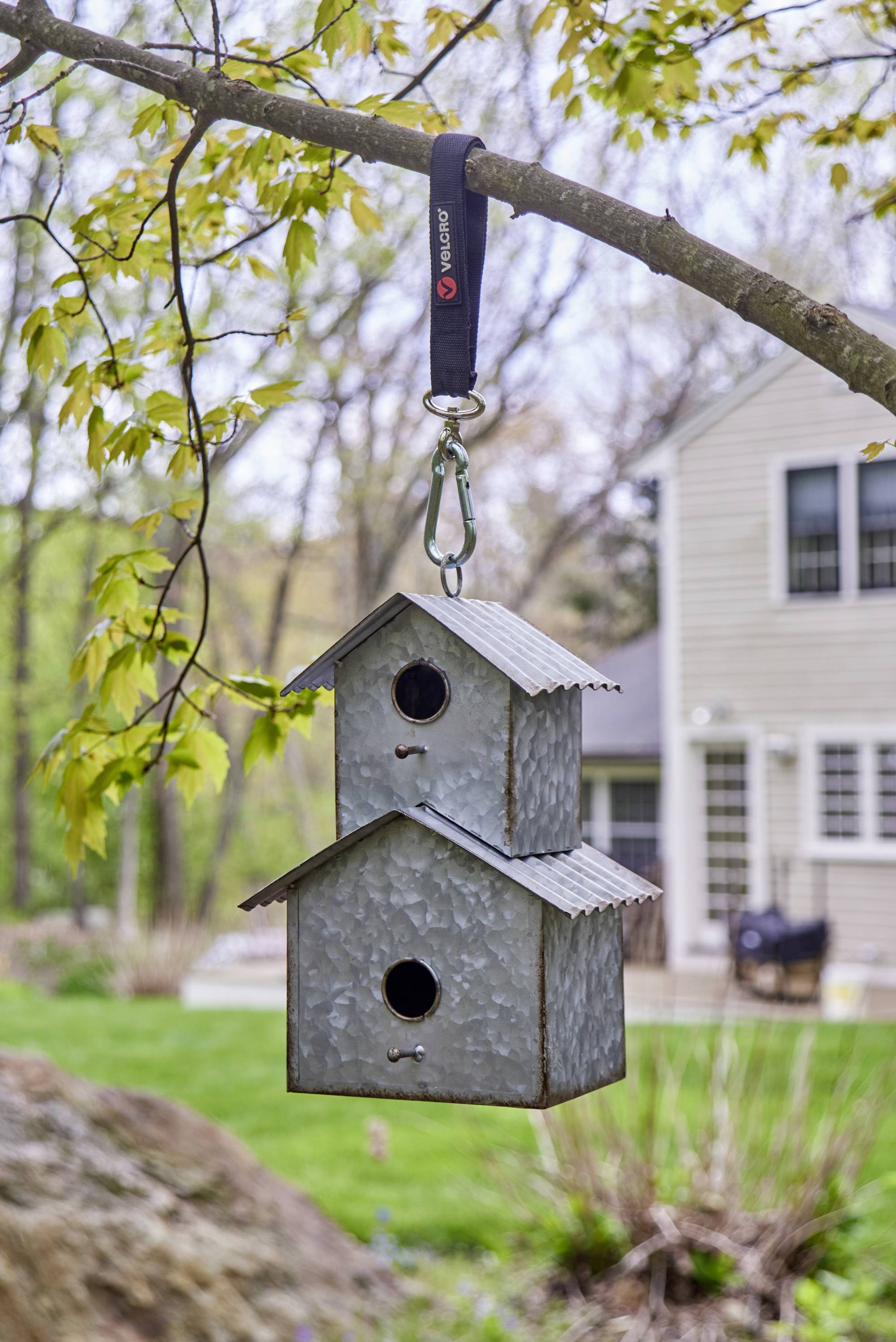 Twee grijze, metalen vogelhuisjes hangen aan een tak. Op de achtergrond is een tuin met een gebouw te zien.