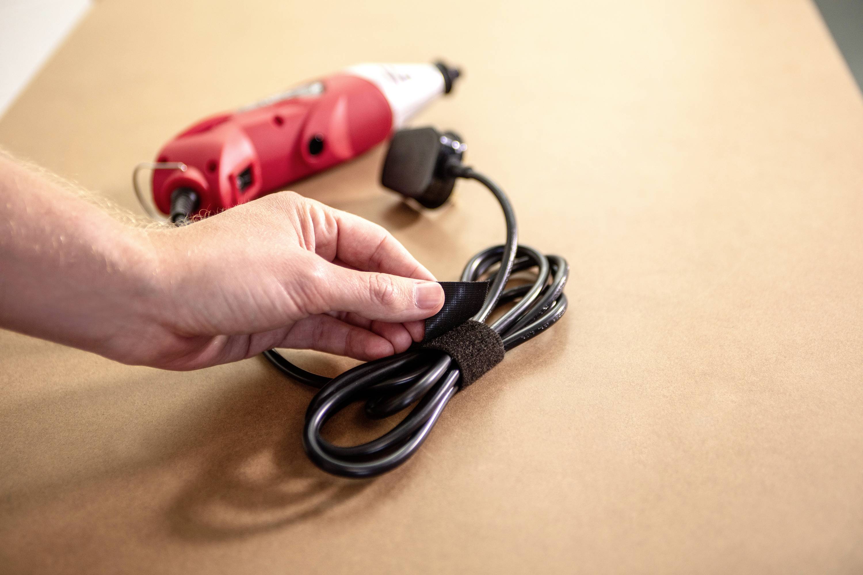 Een hand houdt een zwart kabel met klittenband bij elkaar, op de achtergrond een rood elektrisch gereedschap op een tafel.