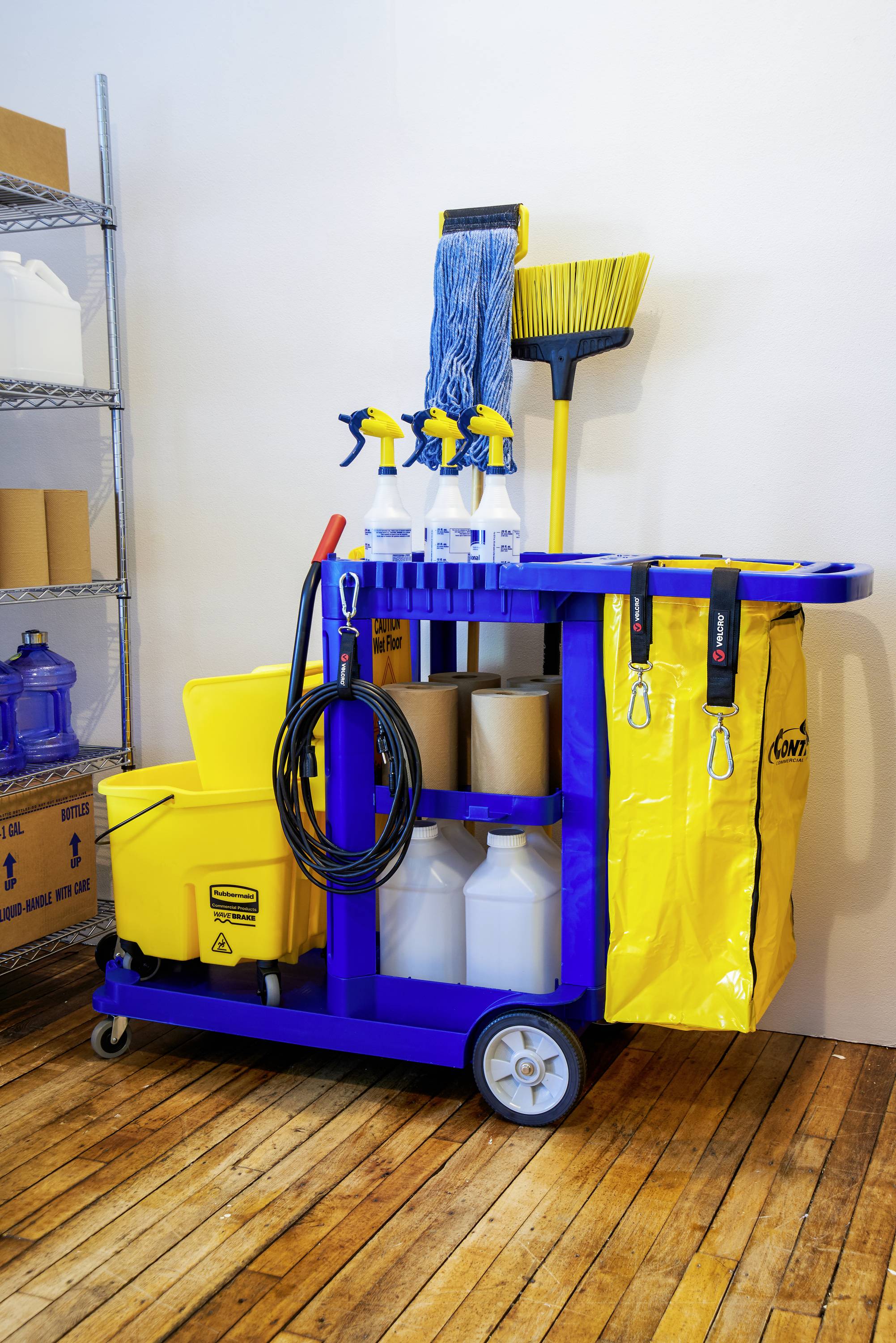 Schoonmaakkar met schoonmaakmiddelen, dweil, bezem en emmer in een opslagruimte op een houten vloer, klaar voor gebouwonderhoud.