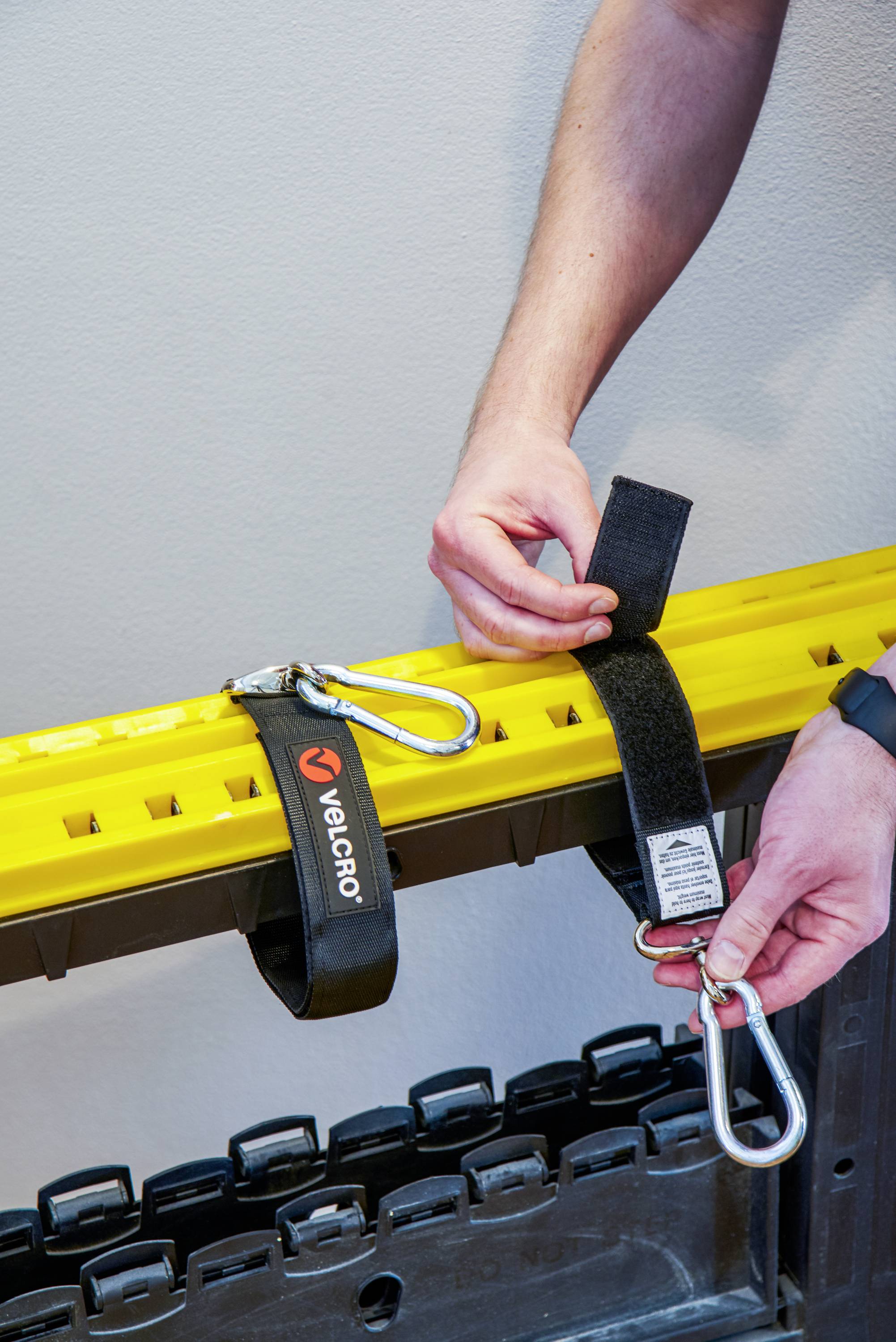 Een persoon bevestigt een gele veiligheidsdrager met zwarte klittenbandsluiting en metalen karabiners aan de muur.