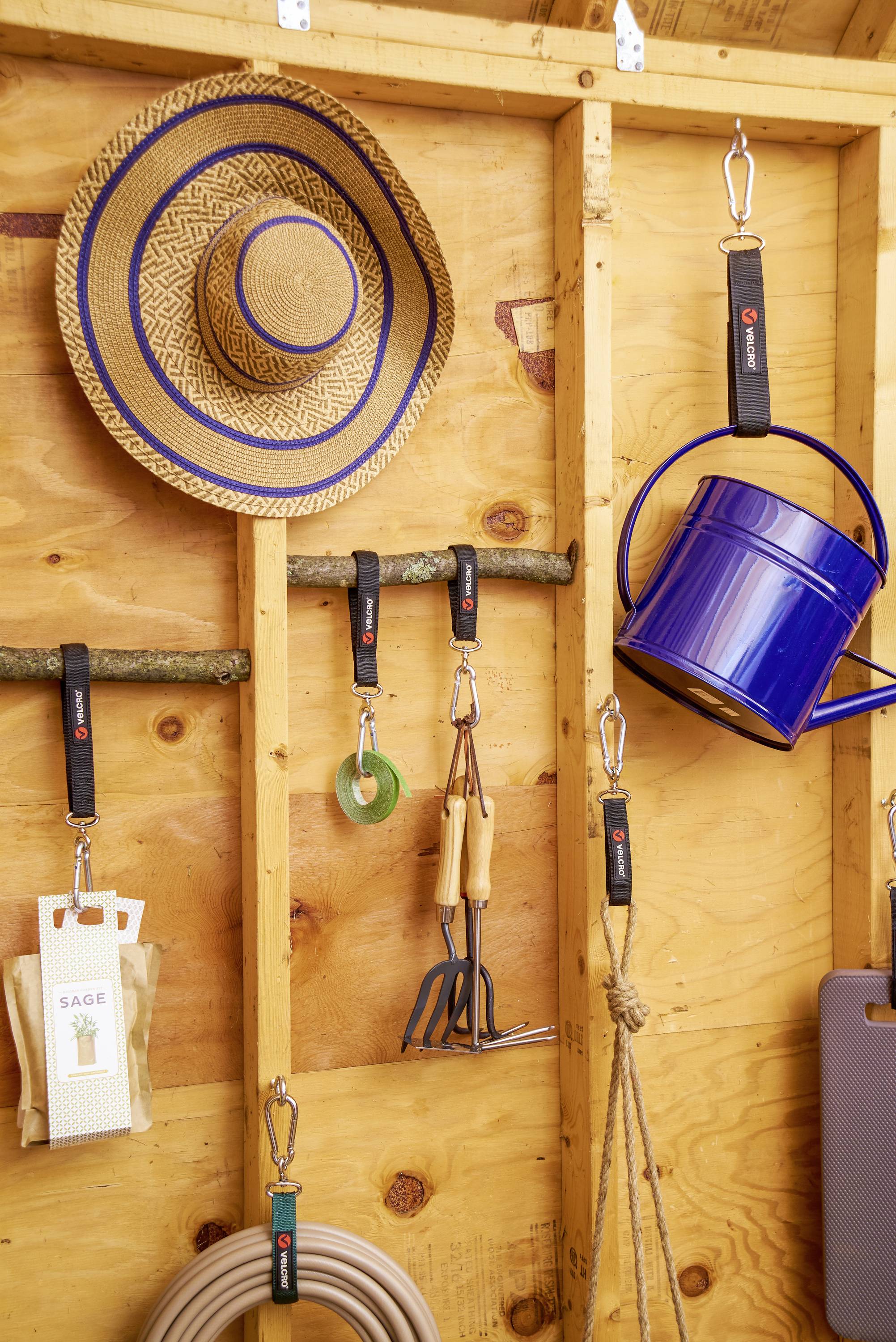 Verschillende tuingereedschappen, een touw en een strohoed hangen aan haken in een houten schuur. Een blauwe emmer is eveneens zichtbaar.