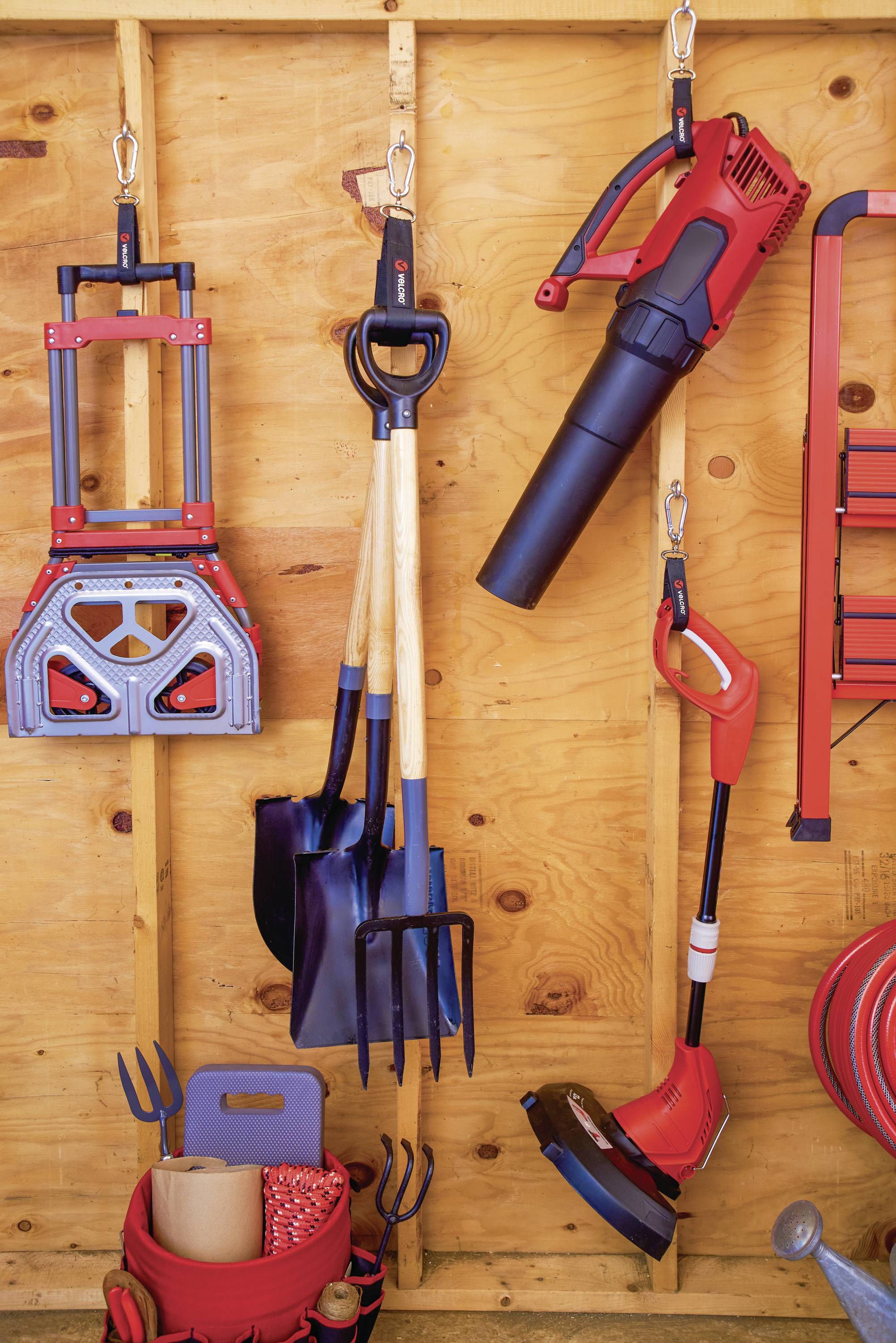 Gereedschappen in een garage hangen aan een houten wand: drie schoppen, een snoeischaar, een bladblazer, een grastrimmer en een gereedschapshouder.