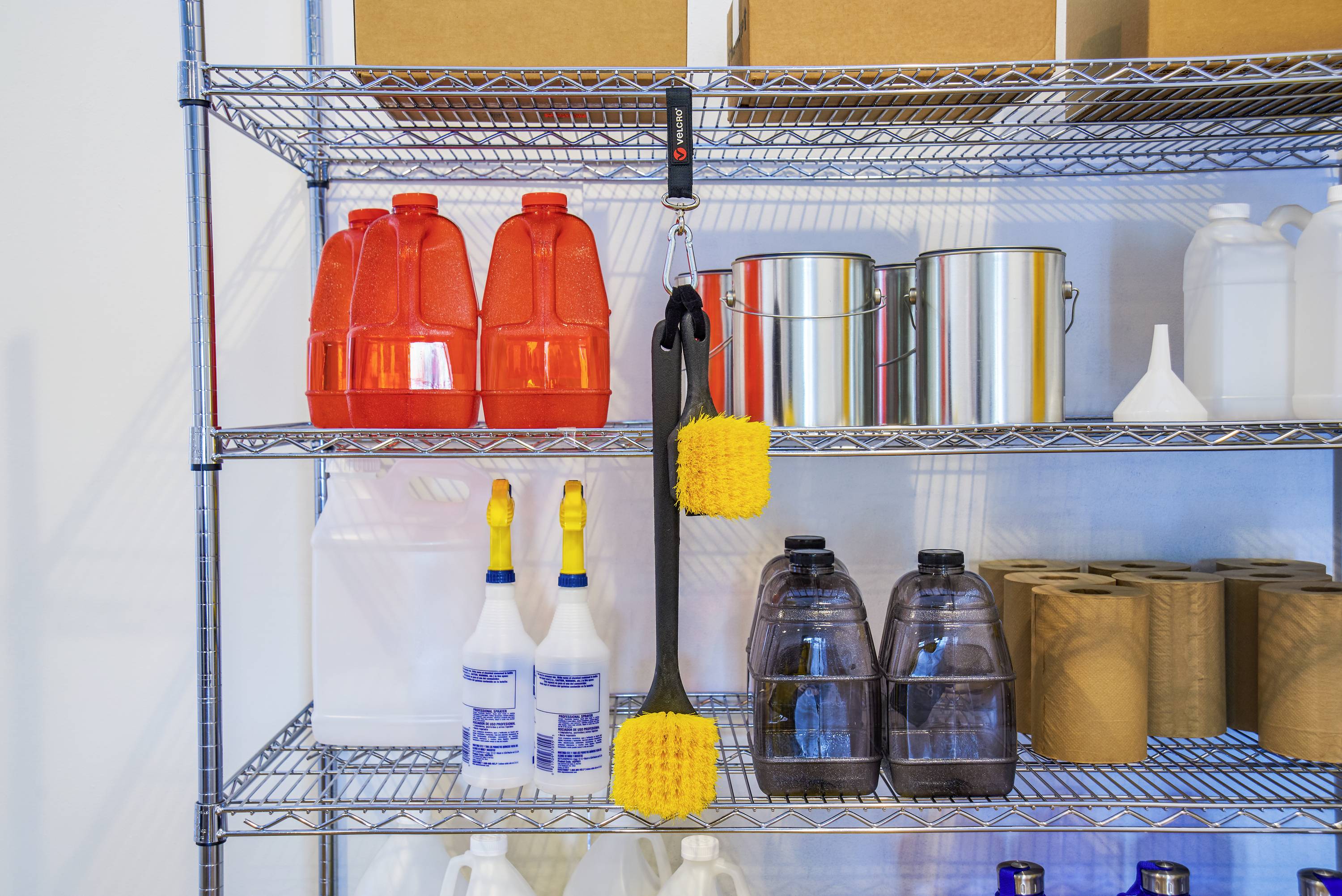 Metalen rek met schoonmaakmiddelen: rode jerrycans, zilveren emmers, spuitflessen, bruine flessen en gele sponsen aan haken.