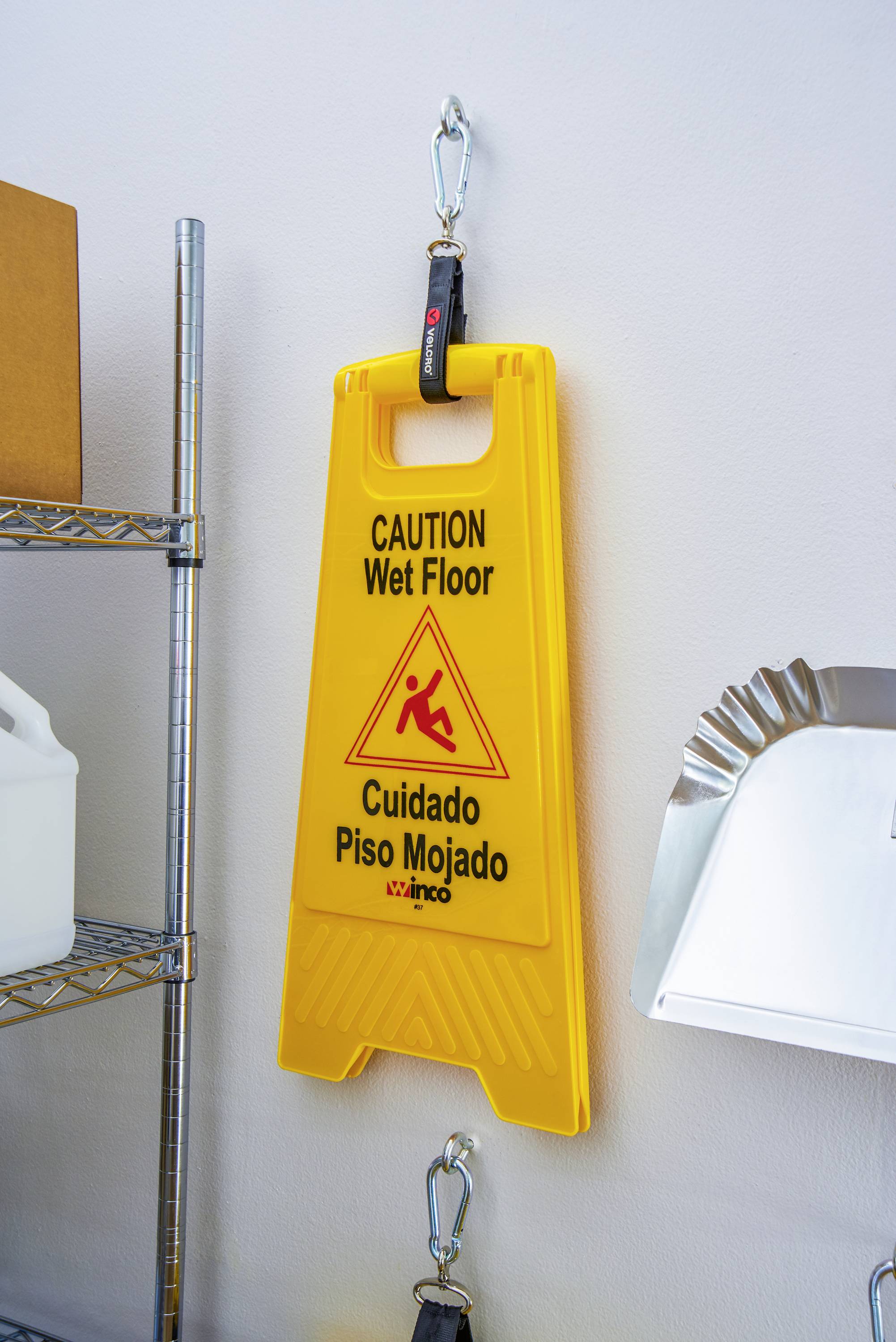 Geel waarschuwingsbord aan de muur met de tekst 'Caution Wet Floor / Cuidado Piso Mojado', naast een rek in een opslagruimte.