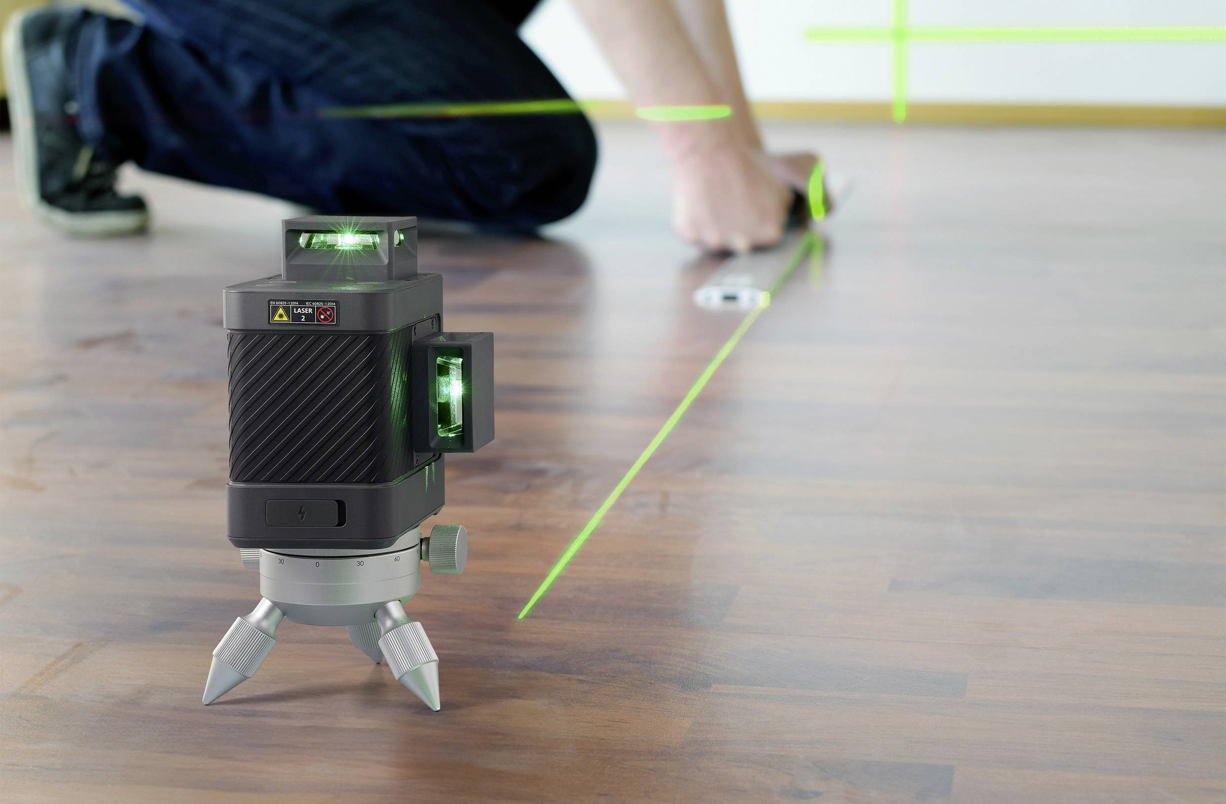 Een groene lasernivelleermachine projecteert lijnen op een houten vloer. Op de achtergrond meet iemand de afstand.