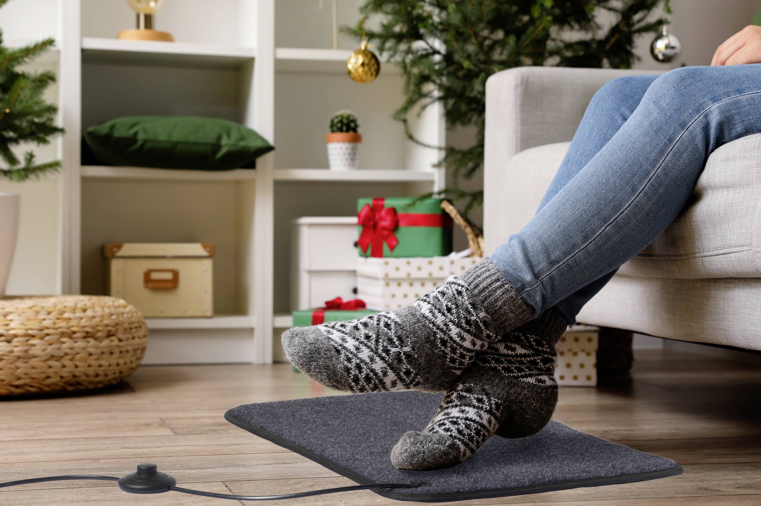 Een persoon in gezellige sokken rust met de voeten op een verwarmde mat, met kerstcadeaus en een versierde kerstboom op de achtergrond, wat een feestelijke sfeer creëert.