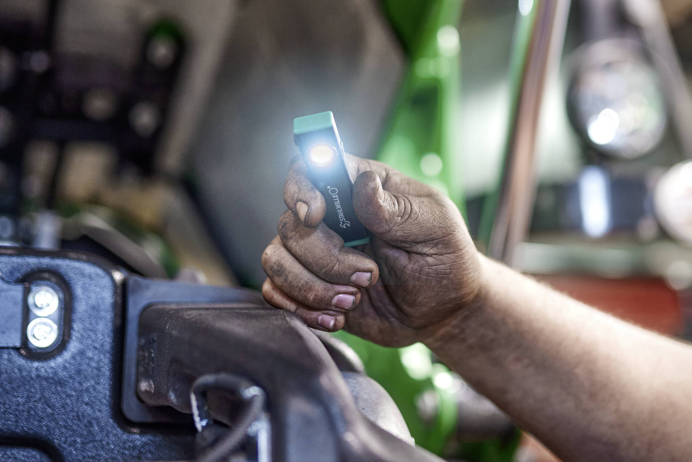 Een hand van een persoon die een kleine zaklamp vasthoudt, waarbij een deel van een machine of voertuig wordt verlicht, wat wijst op reparatie- of onderhoudswerk.