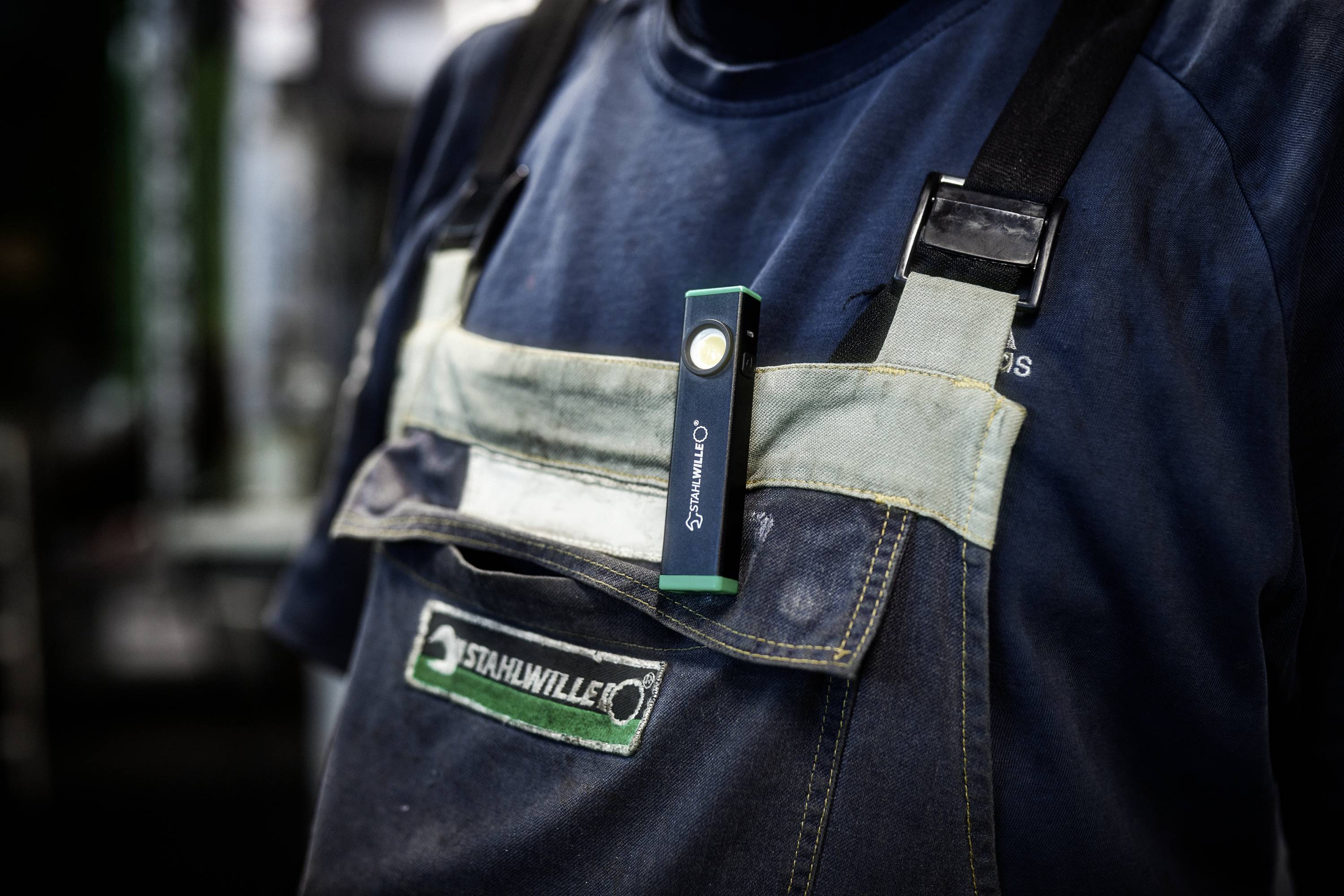 Een persoon die blauwe overalls draagt met een 'STAHLWILLE' badge en een groene penlight vastgeklikt aan de voorste zak.