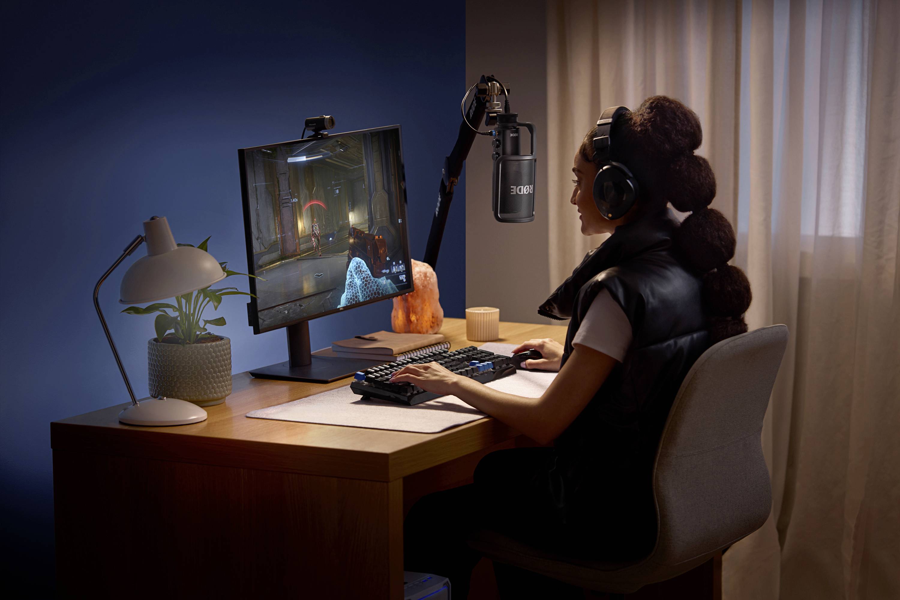 Een persoon zit aan een bureau, speelt een videospel op een scherm, draagt koptelefoon en spreekt in een microfoon.