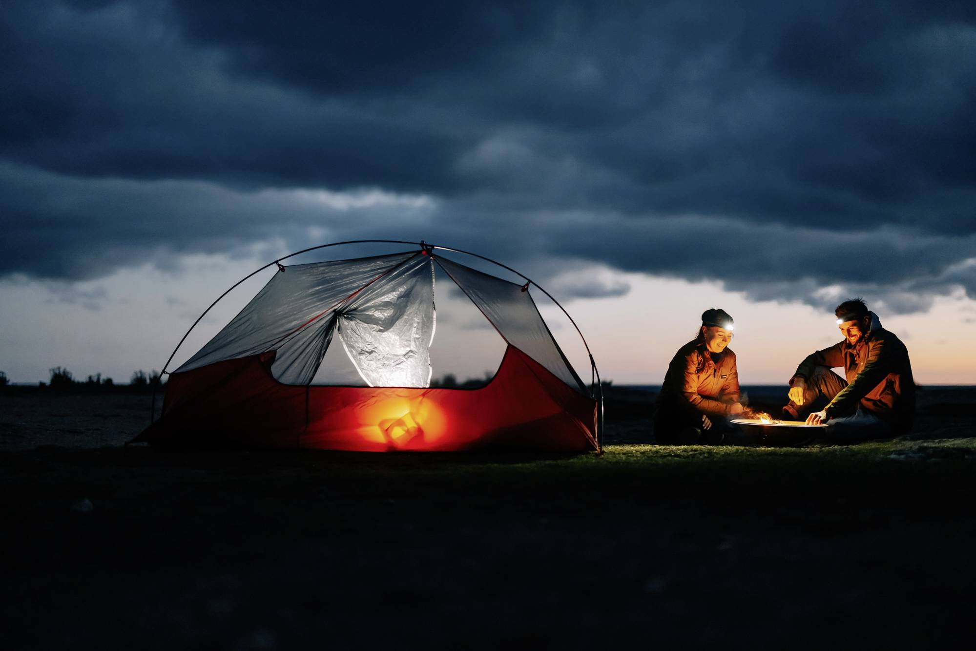 Twee personen zitten 's avonds bij een kampvuur naast een verlichte tent. Donkere lucht en een rustig landschap op de achtergrond.